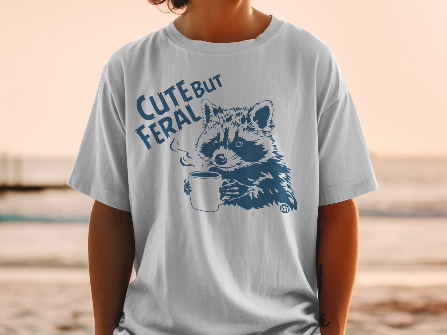 White t-shirt with dark blue graphic of raccoon holding coffee cup and text "CUTE BUT FERAL".