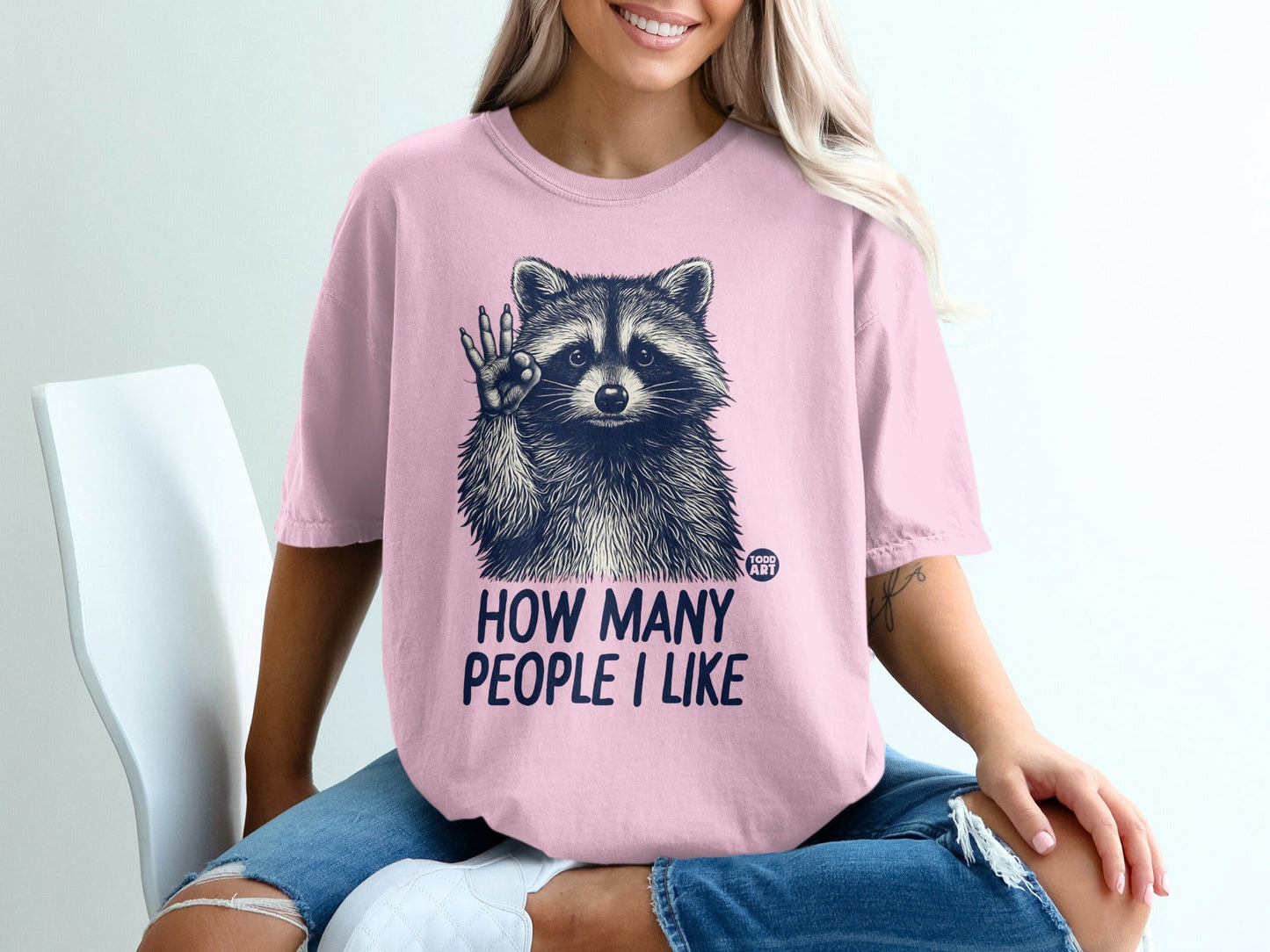 Pink t-shirt with raccoon graphic and text "HOW MANY PEOPLE I LIKE".