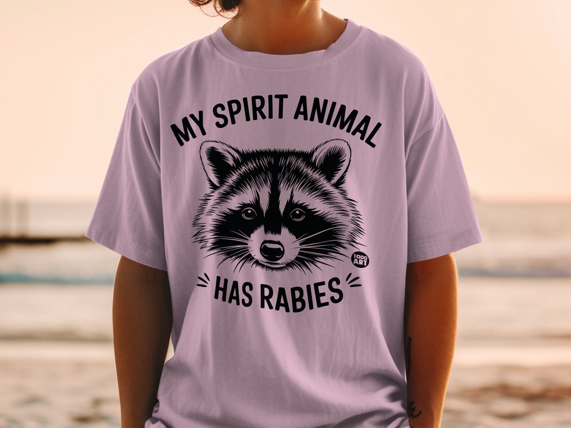 Lavender t-shirt with raccoon graphic and text "MY SPIRIT ANIMAL HAS RABIES".