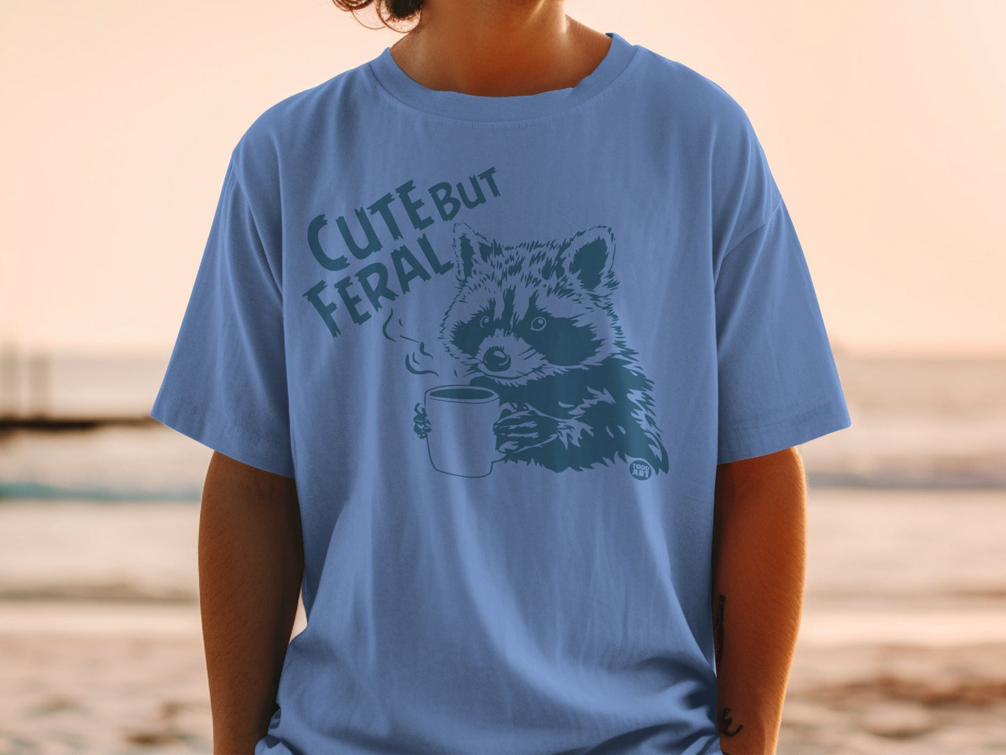 Blue t-shirt with raccoon holding coffee mug, "CUTE BUT FERAL" text.