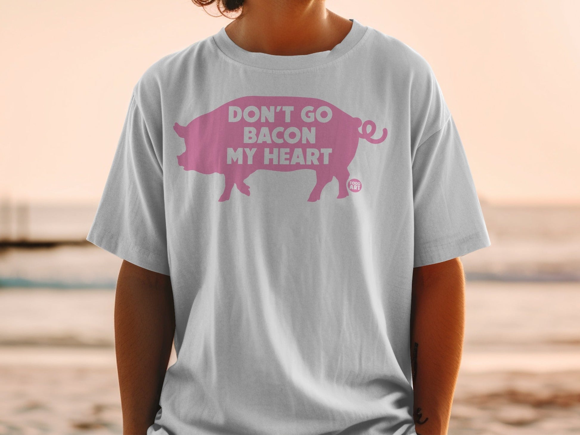 White t-shirt featuring a pink pig silhouette with text "DON'T GO BACON MY HEART".