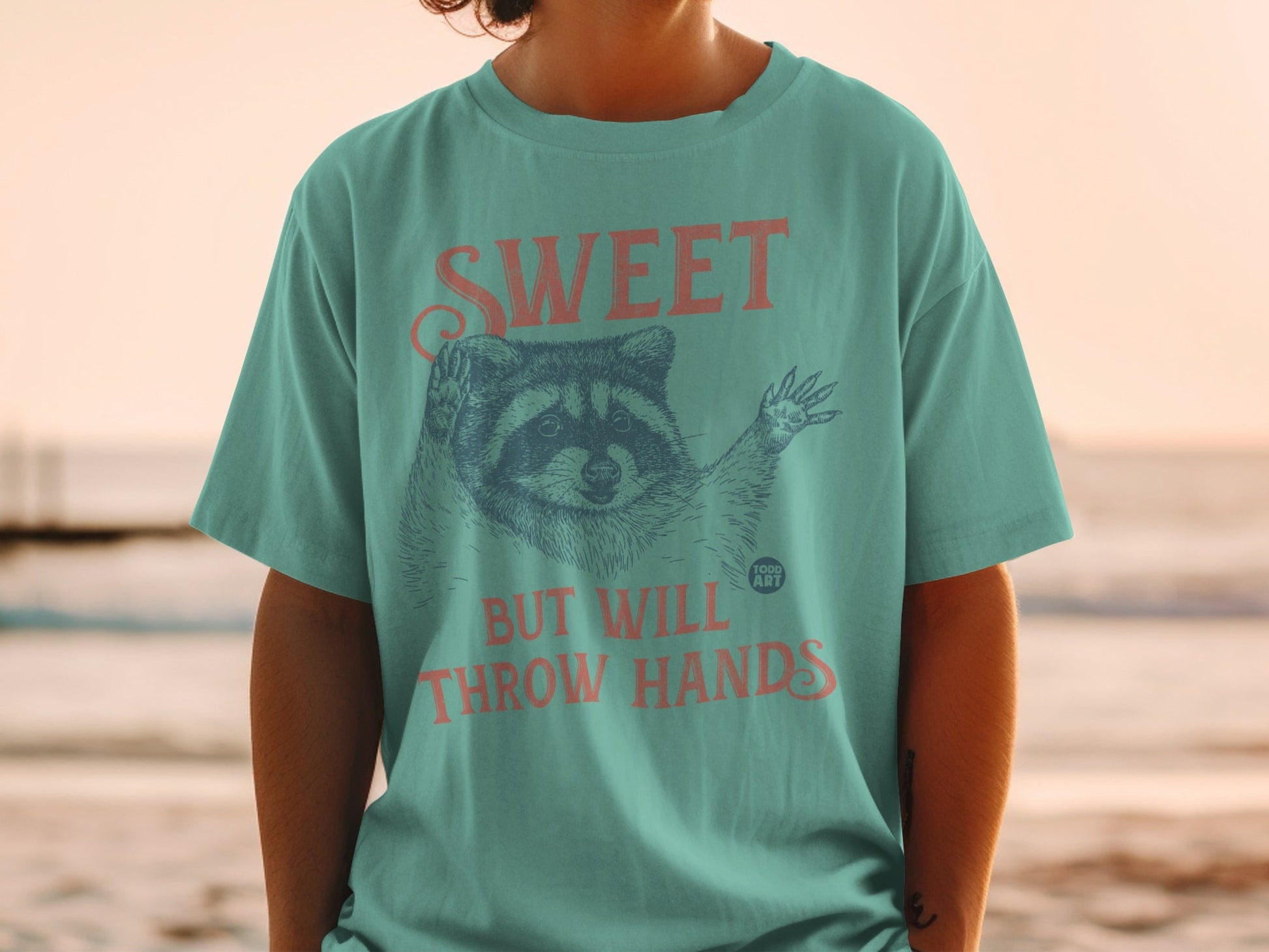 Teal t-shirt with raccoon graphic and "SWEET BUT WILL THROW HANDS" text.