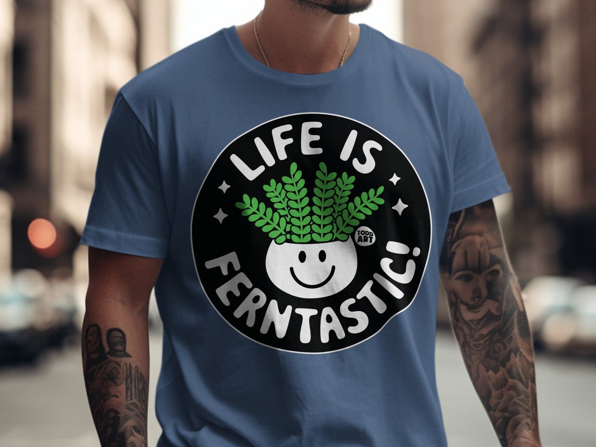 Blue t-shirt with a circular graphic featuring a smiling fern in a pot that reads "LIFE IS FERNTASITC!"