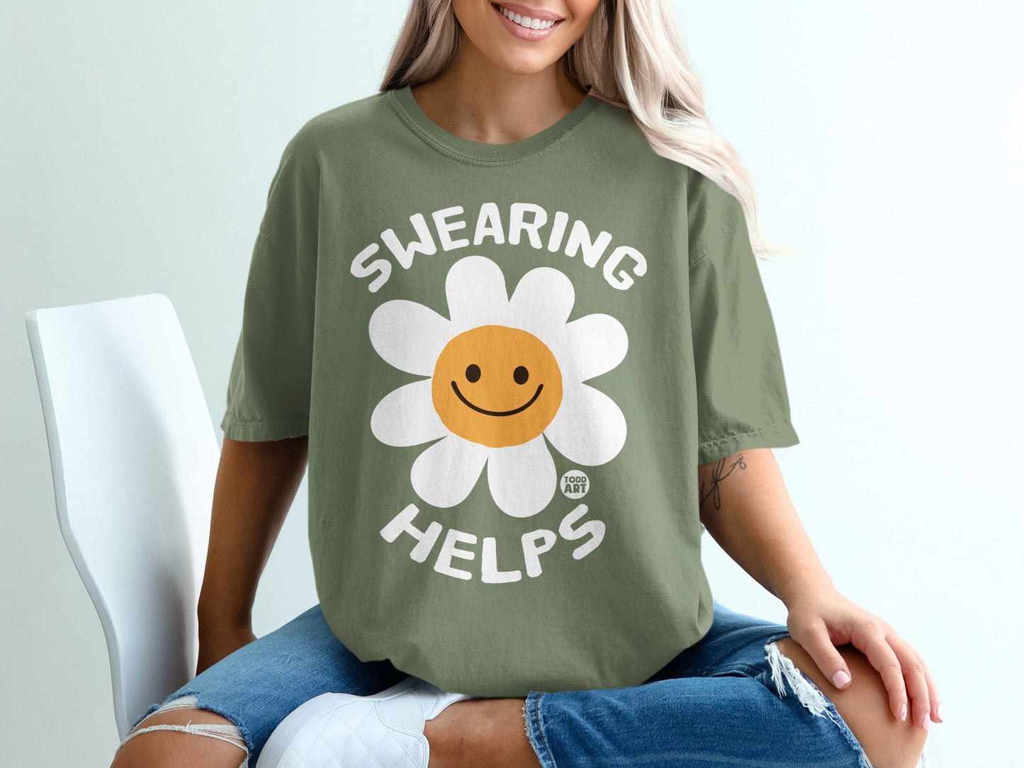 Olive green t-shirt with a smiling flower graphic and text "SWEARING HELPS".