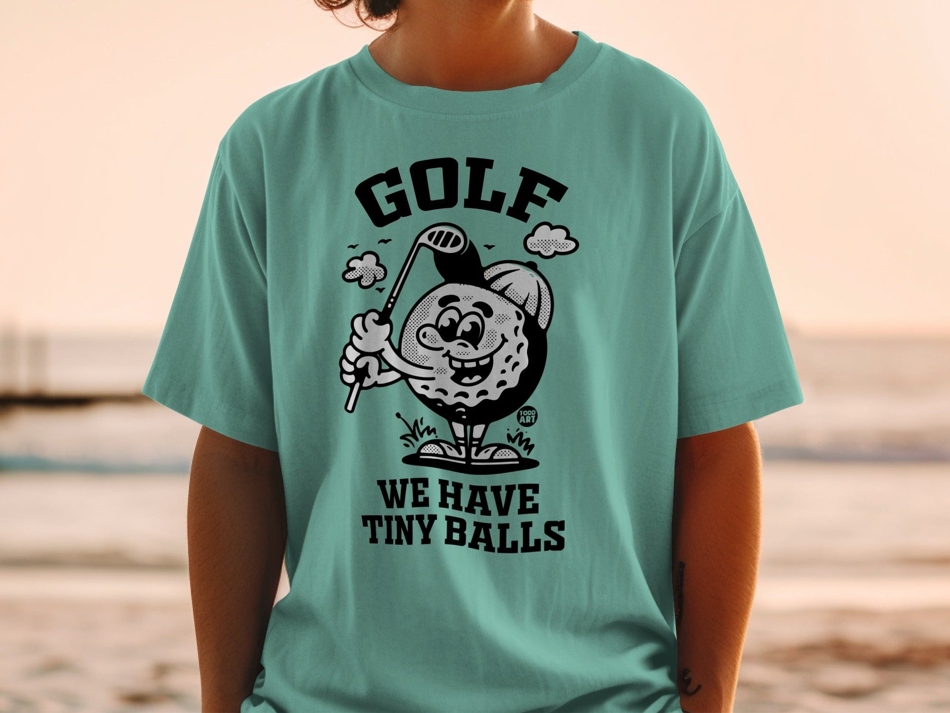 T-shirt featuring a cartoon golf ball character swinging a club with the text "WE HAVE TINY BALLS."