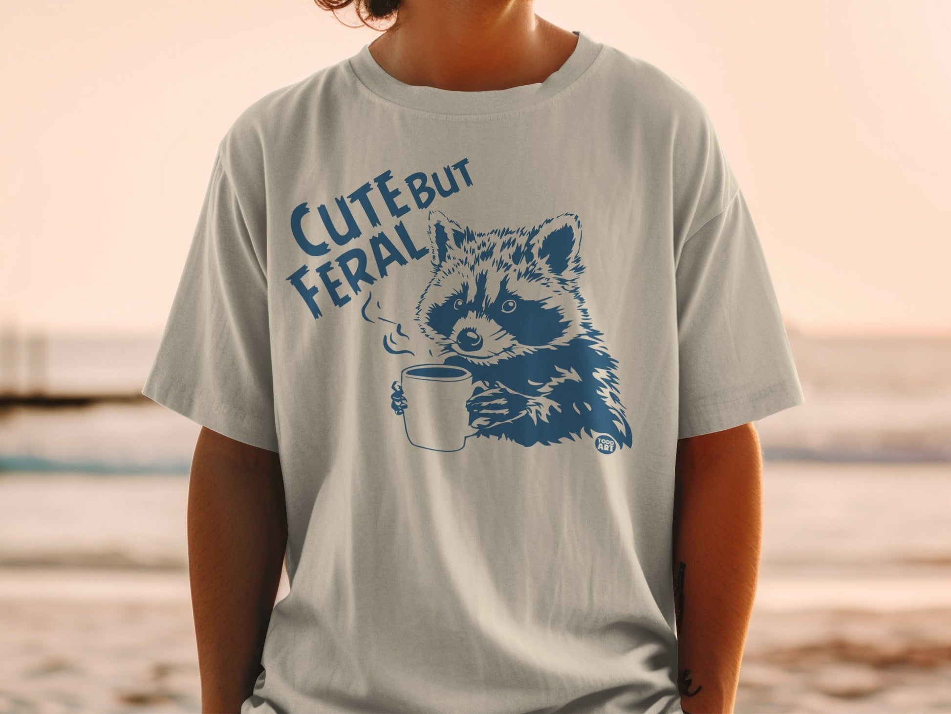 T-shirt with raccoon holding coffee cup and text "CUTE BUT FERAL".