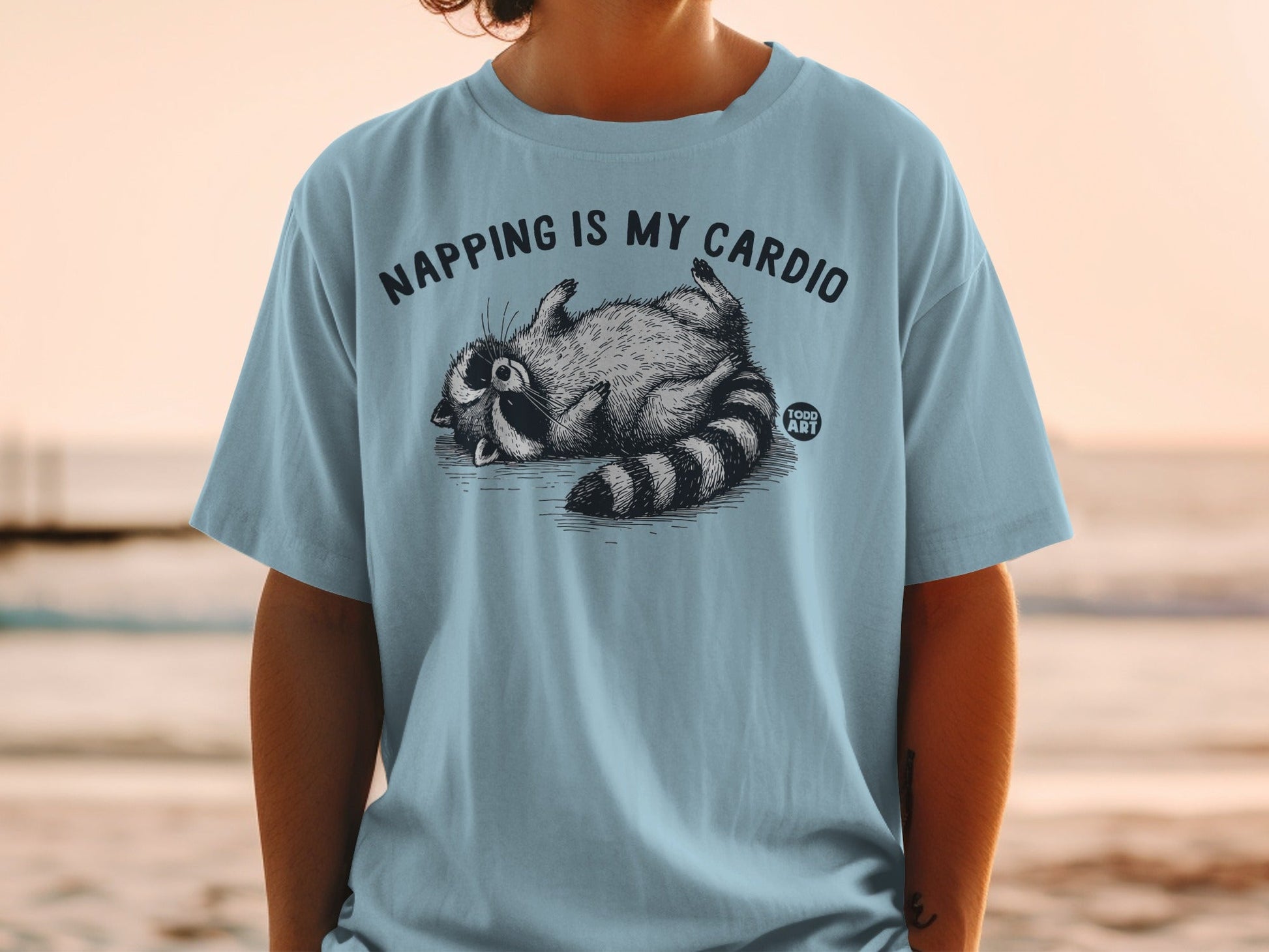 T-shirt with a raccoon lying on its back, text "NAPPING IS MY CARDIO" above.