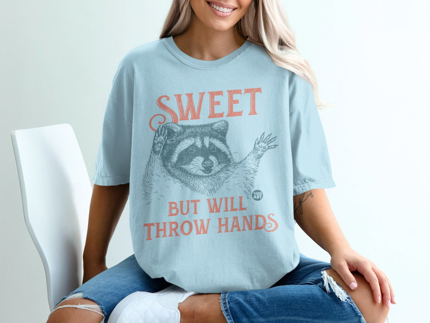 Light blue t-shirt with raccoon graphic and text "SWEET BUT WILL THROW HANDS".