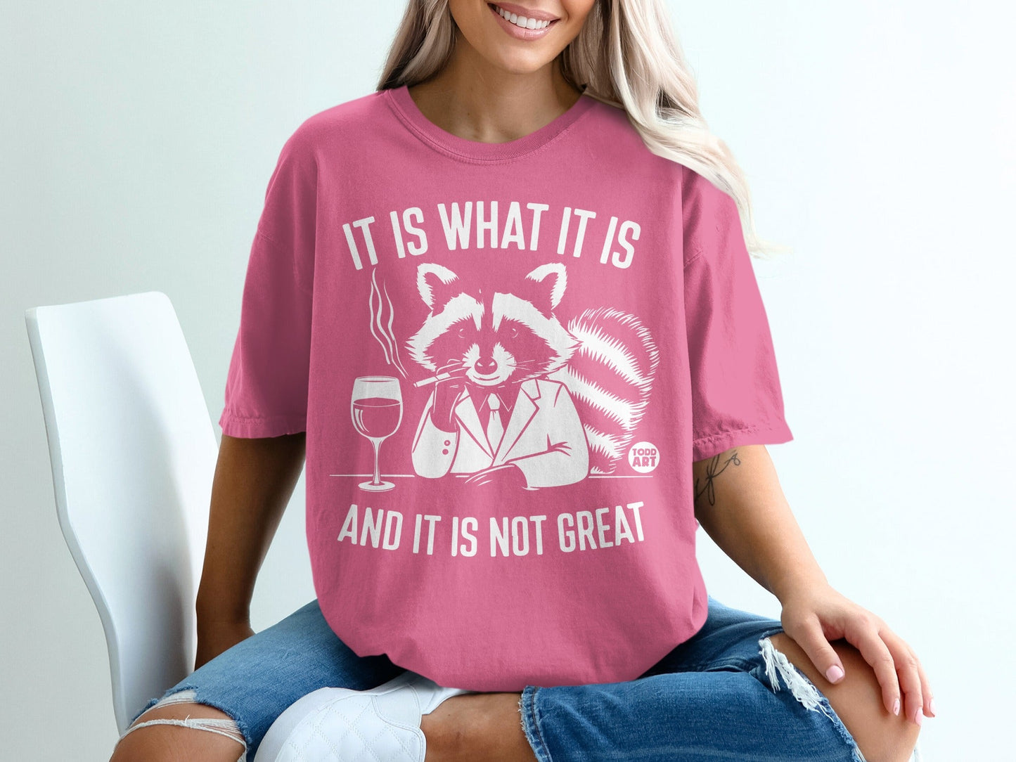 Pink t-shirt with raccoon smoking and drinking wine, text: "It is what it is and it is not great".