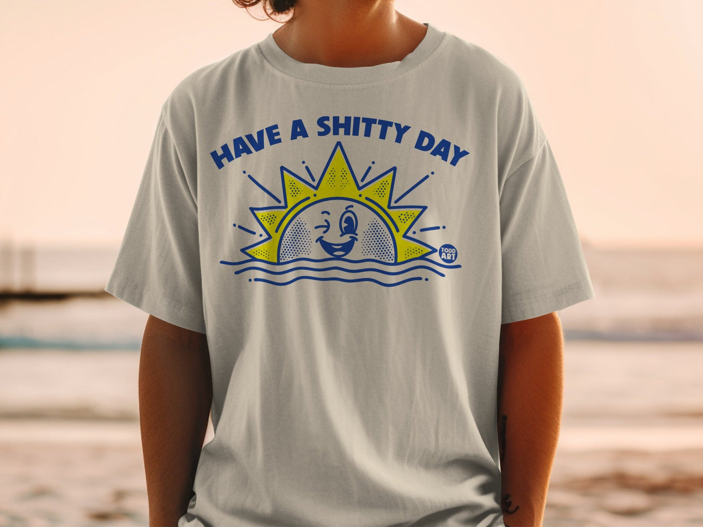 T-shirt with a cartoon sun saying "HAVE A SHITTY DAY" above wavy blue lines.
