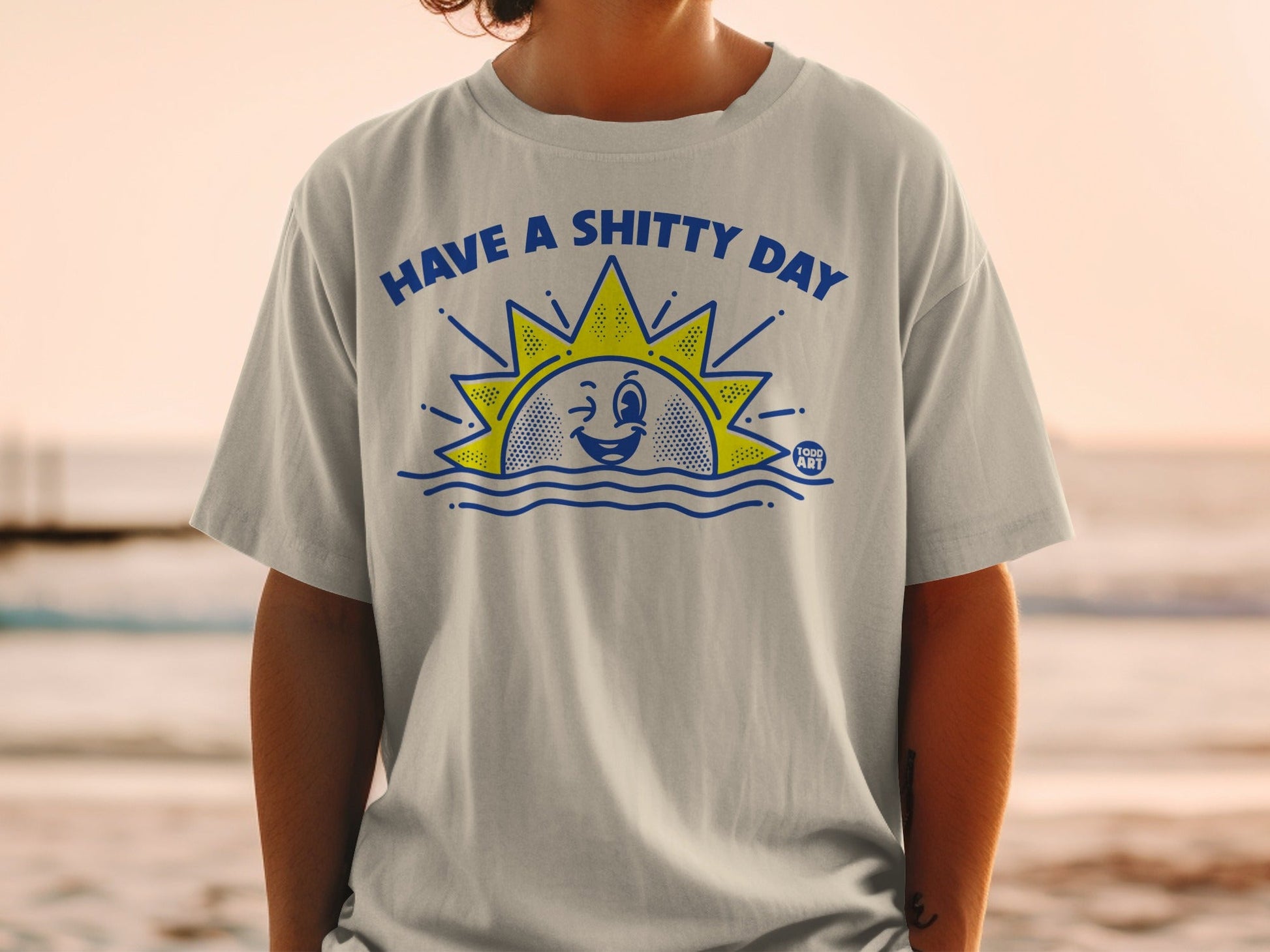 T-shirt with a cartoon sun saying "HAVE A SHITTY DAY" above wavy blue lines.