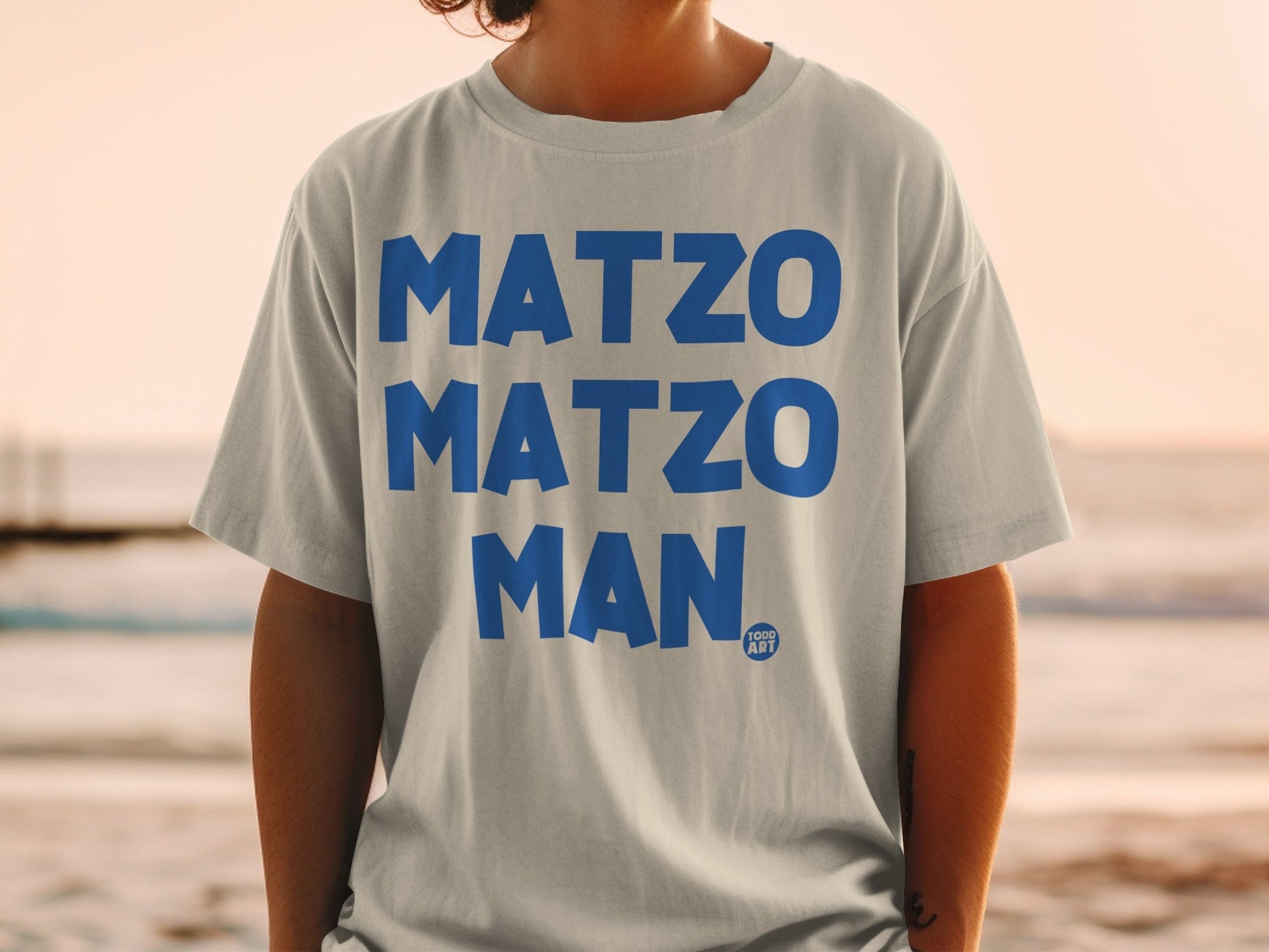 Light grey t-shirt with bold blue "MATZO MATZO MAN" text and a small "100G ART" logo.