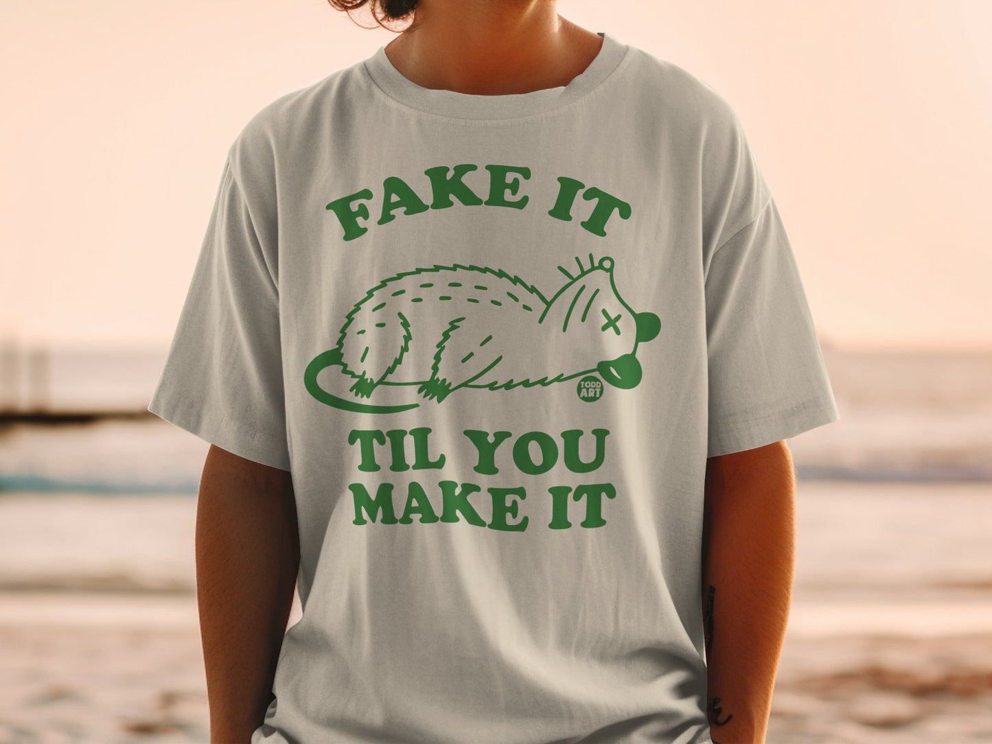 Light grey t-shirt with green text "FAKE IT TIL YOU MAKE IT" and a cartoon possum.