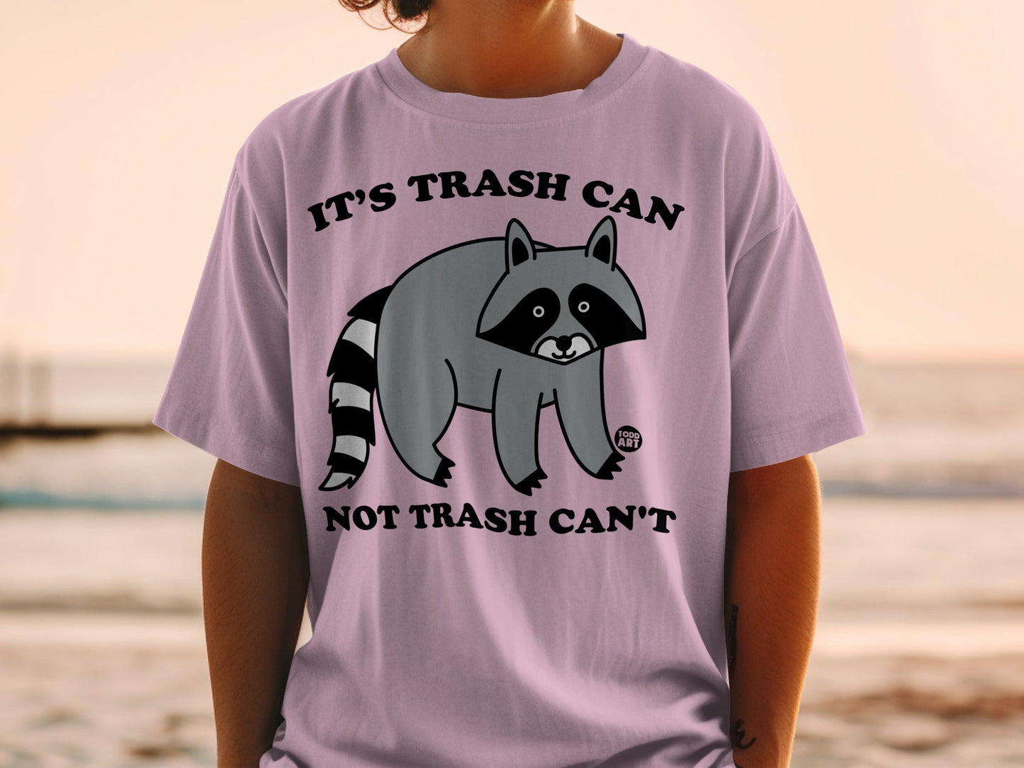 Light purple t-shirt with a raccoon graphic and text "IT'S TRASH CAN NOT TRASH CAN'T".