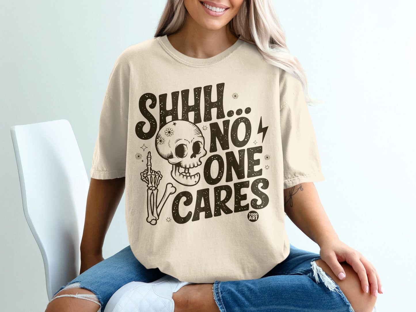 Cream t-shirt with skeleton, middle finger, and "SHHH... NO ONE CARES" text.