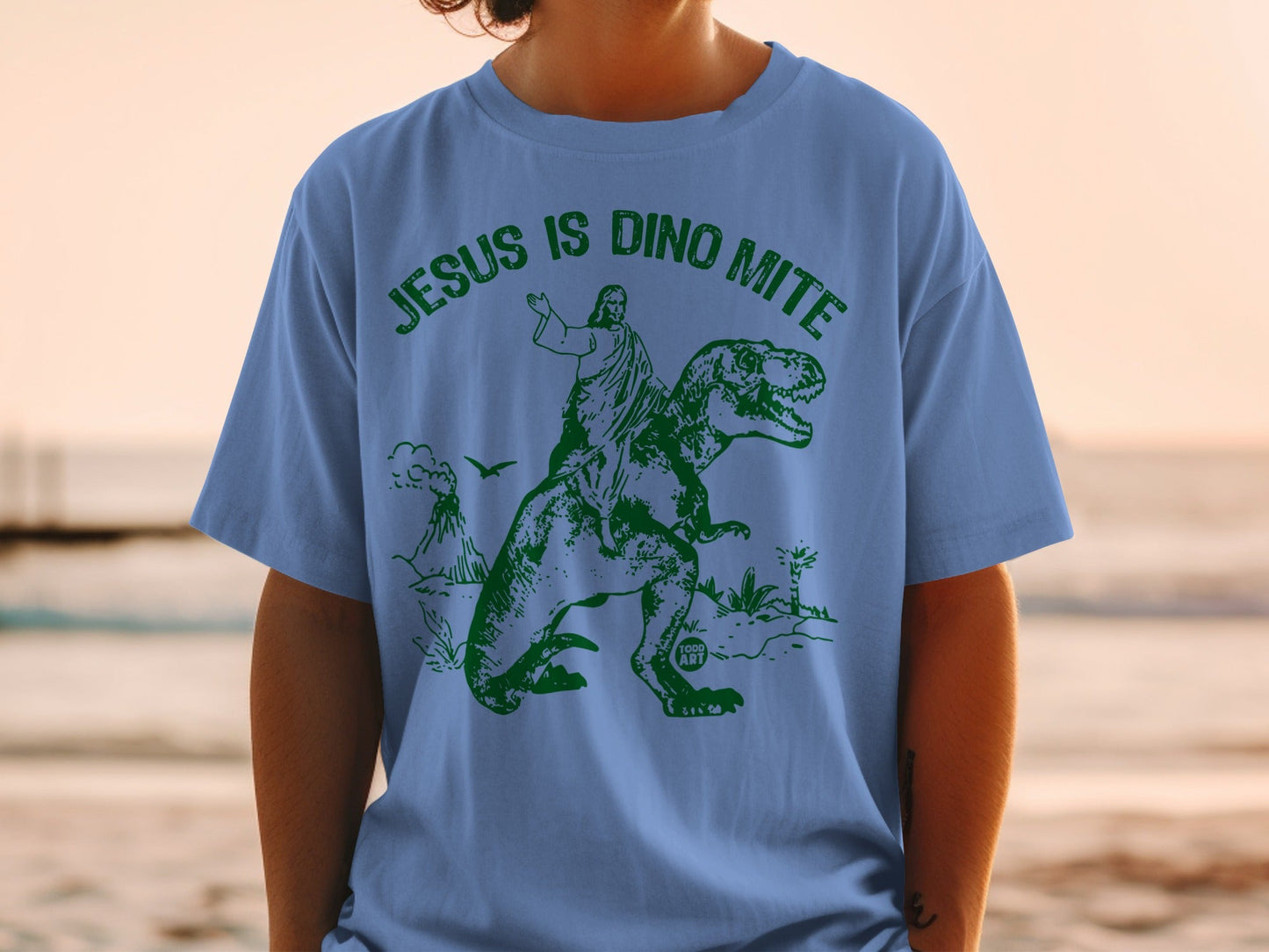 Blue t-shirt with green graphic of Jesus riding a dinosaur.