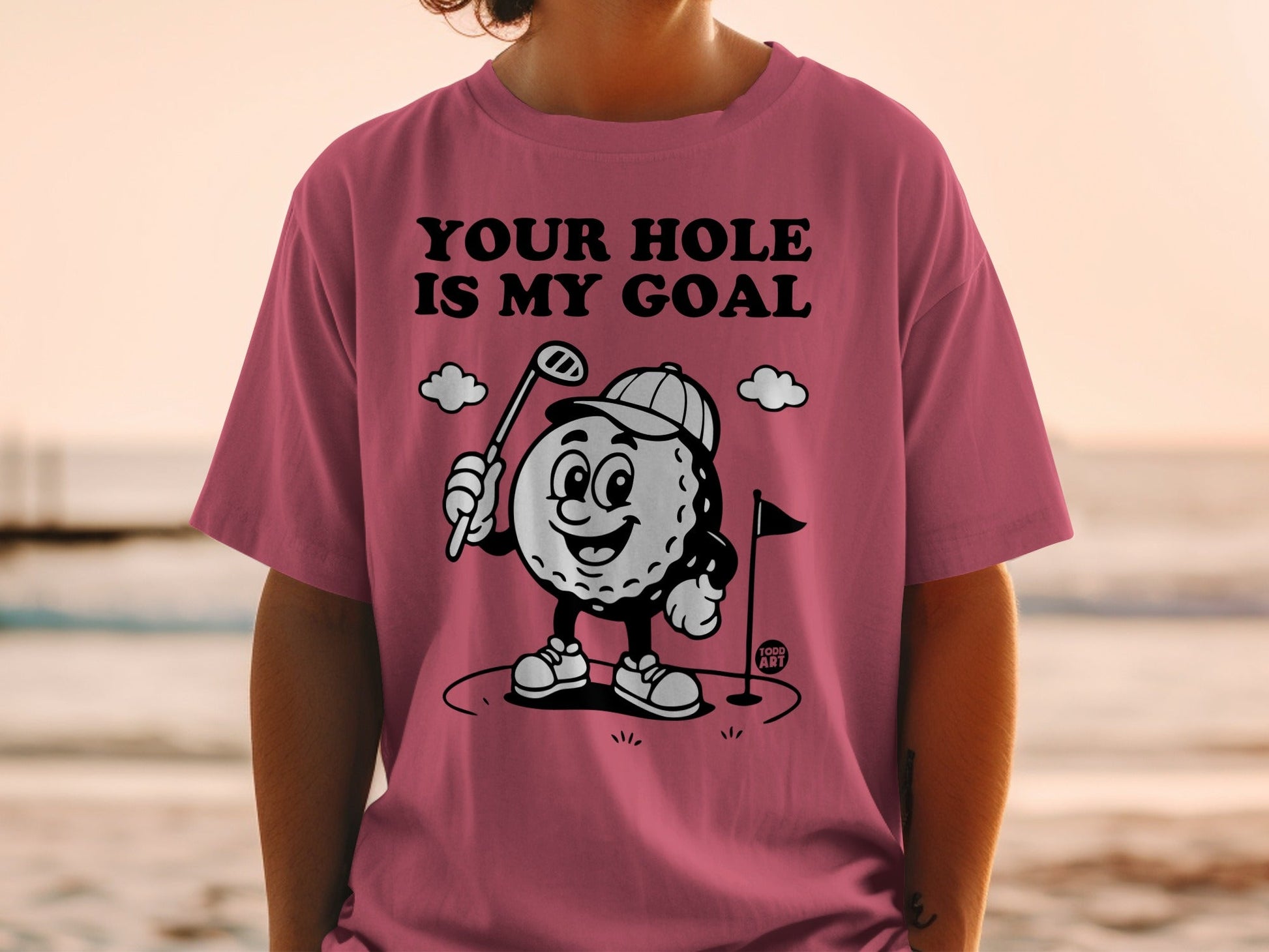 T-shirt with cartoon golf ball saying "YOUR HOLE IS MY GOAL" on a beach background.