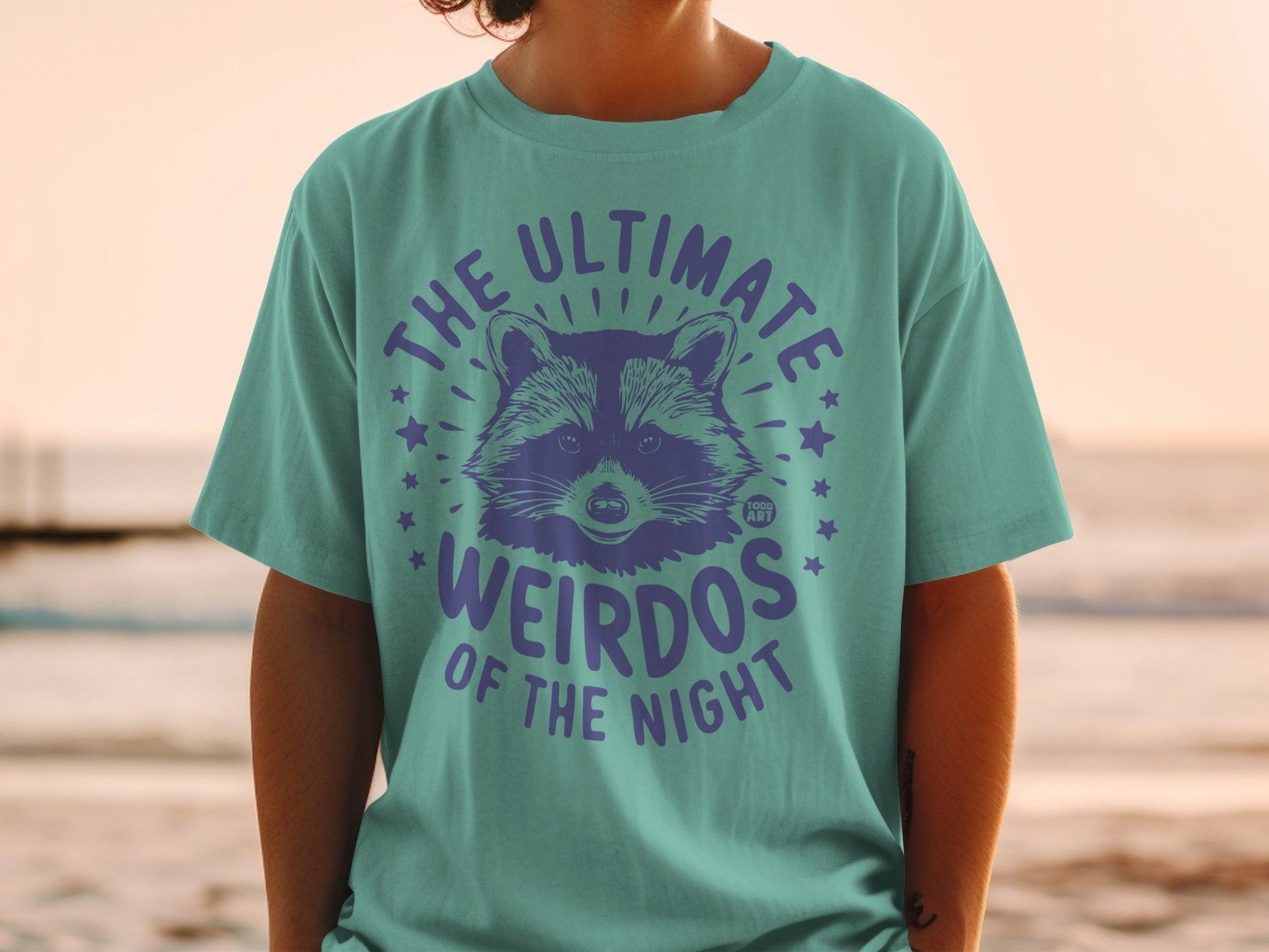 Teal t-shirt with purple raccoon graphic and "The Ultimate Weirdos of the Night" text.