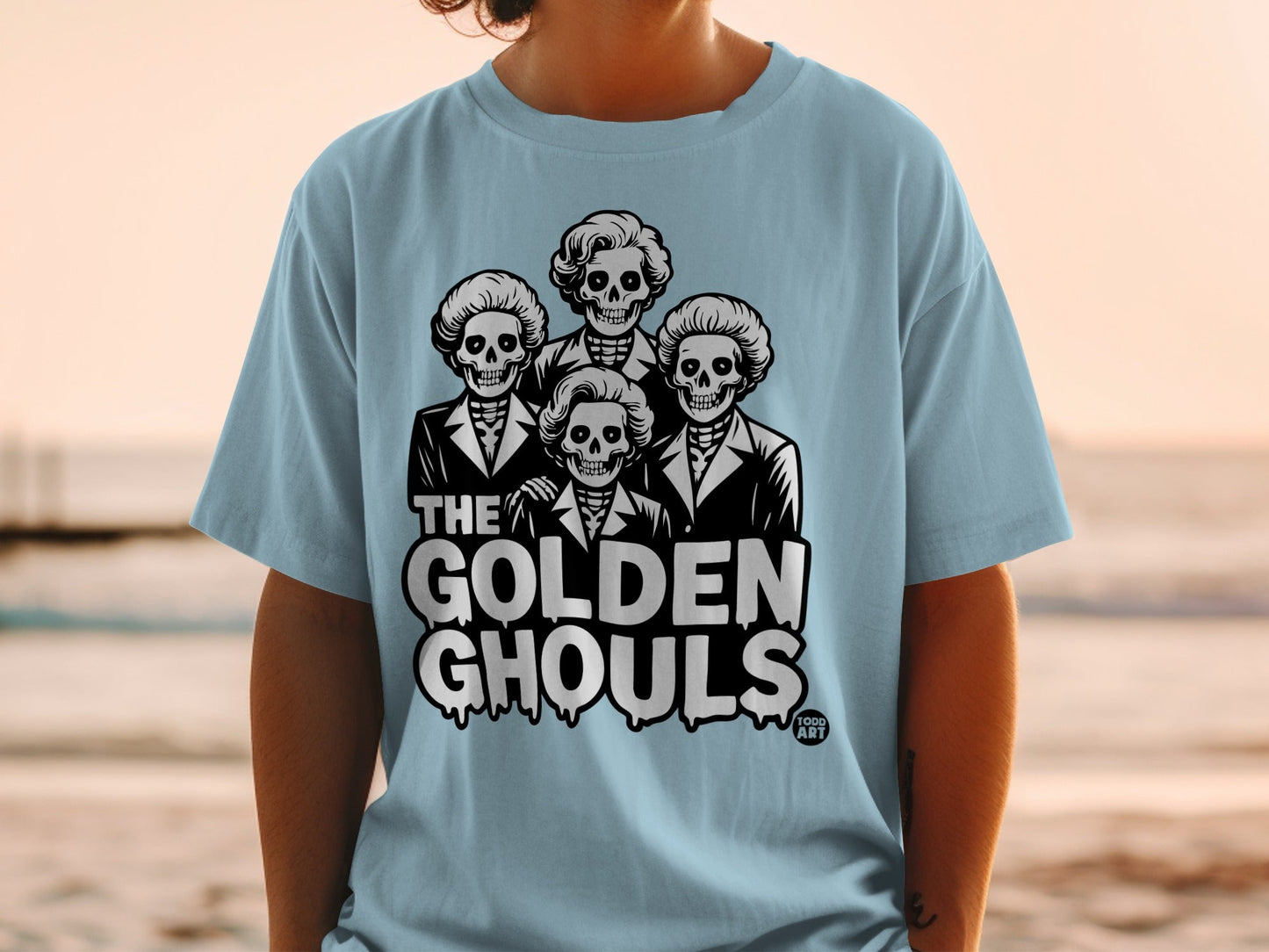 Light blue t-shirt featuring the text "THE GOLDEN GHOULS" above four skeleton heads.