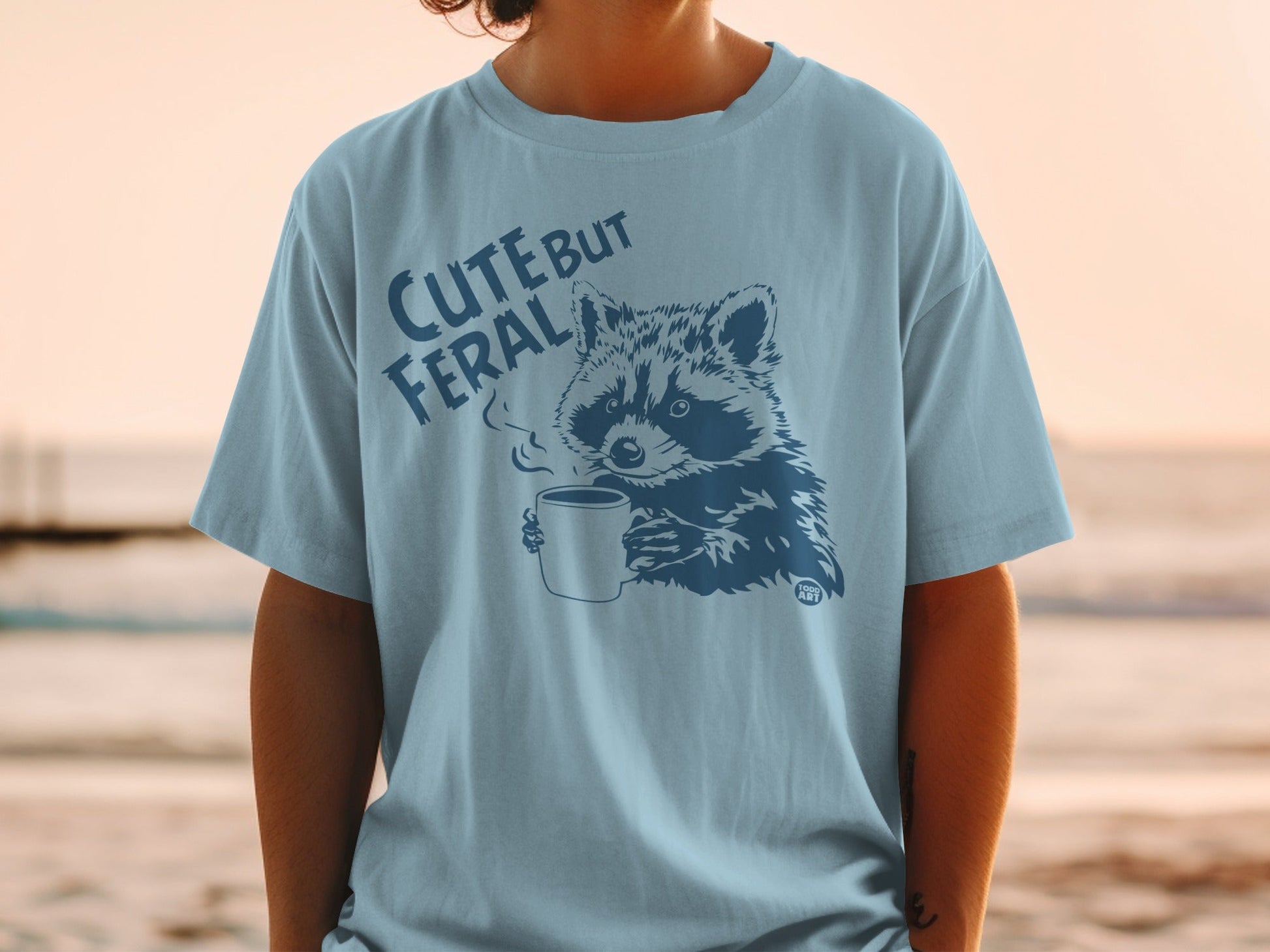 T-shirt with dark blue graphic of raccoon holding coffee cup, text "CUTE BUT FERAL".