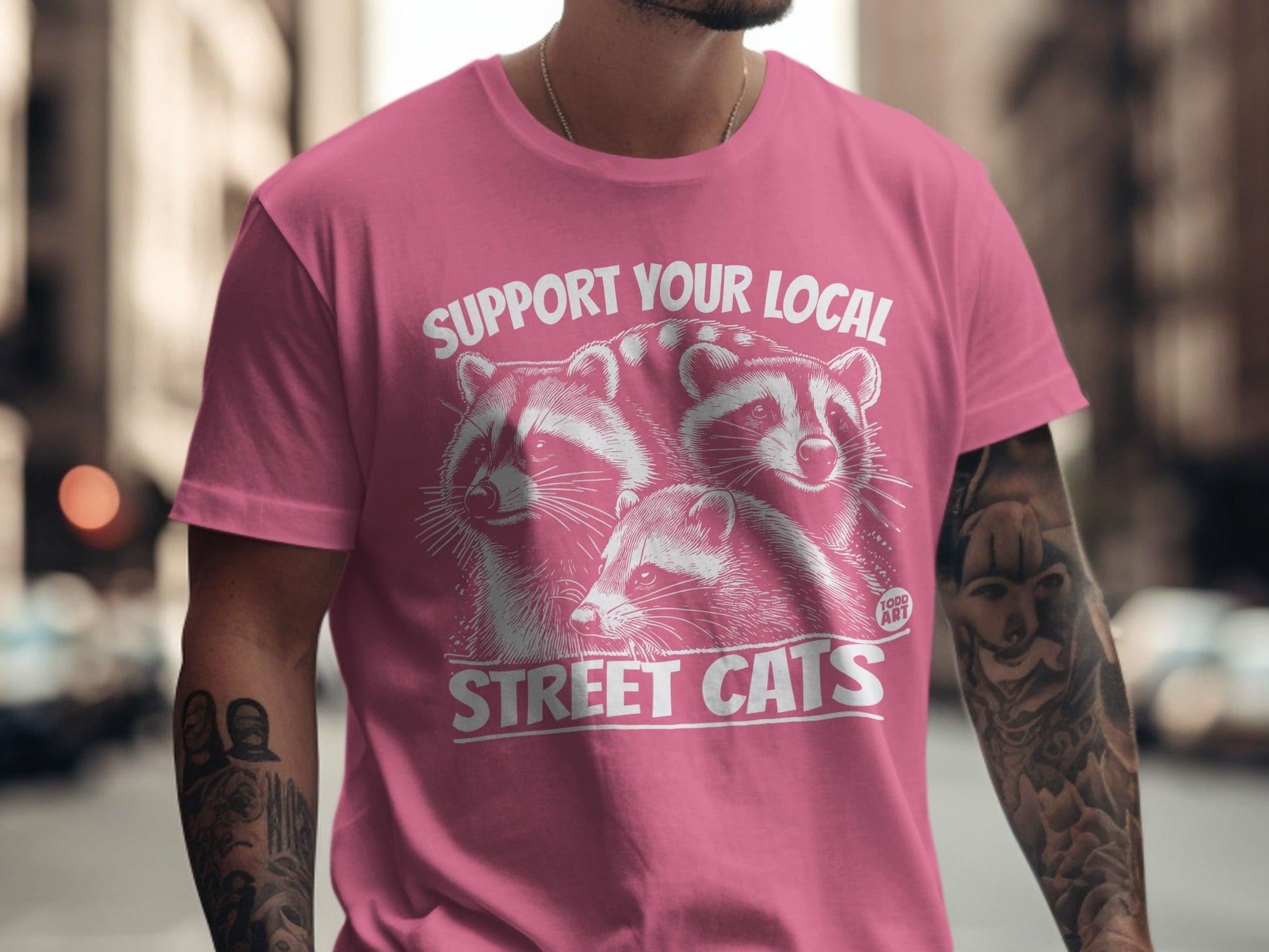 Pink t-shirt with graphic of three raccoons and text "SUPPORT YOUR LOCAL STREET CATS".