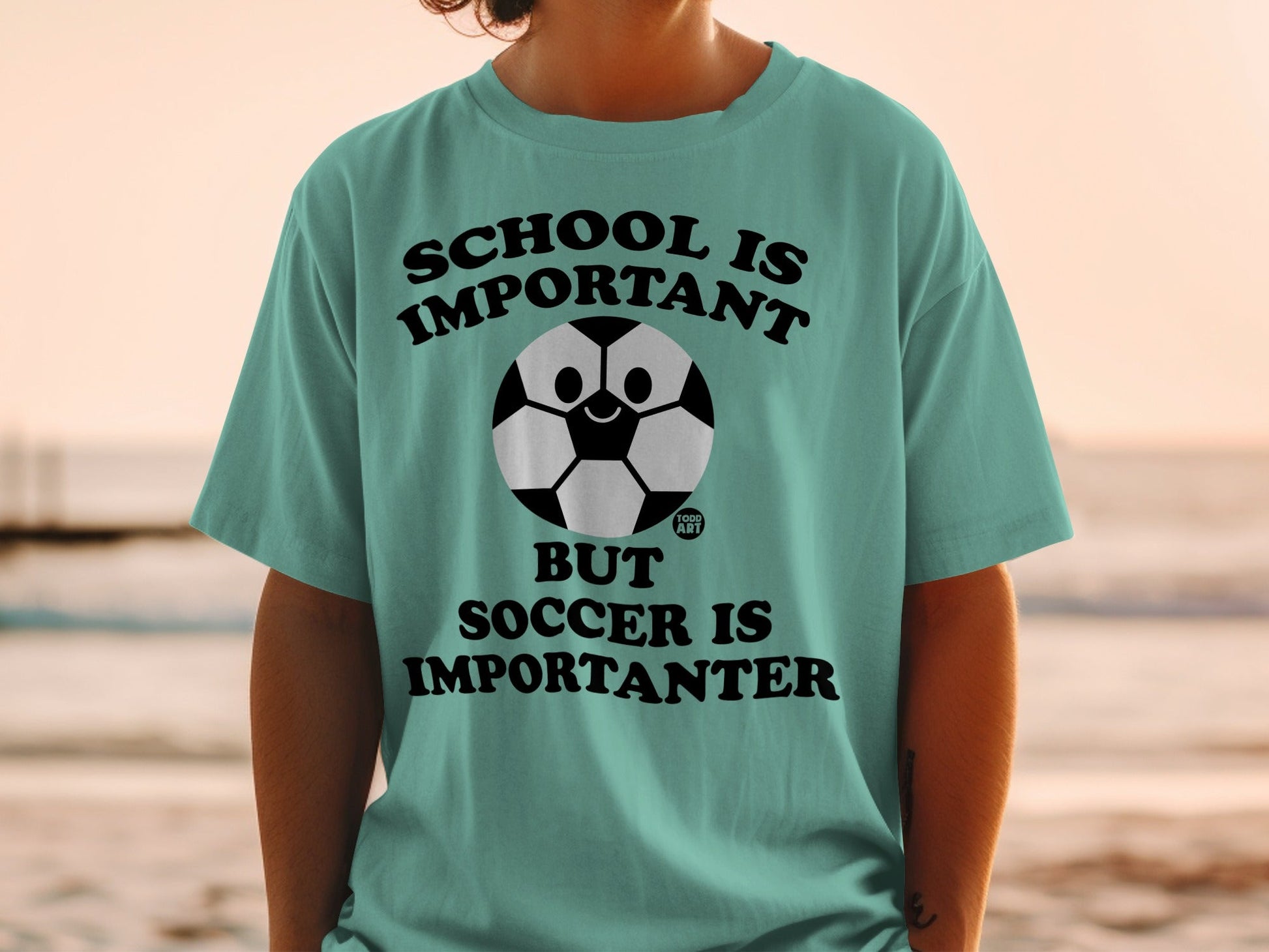 Teal t-shirt with cartoon soccer ball and text: School is important, but soccer is importanter.