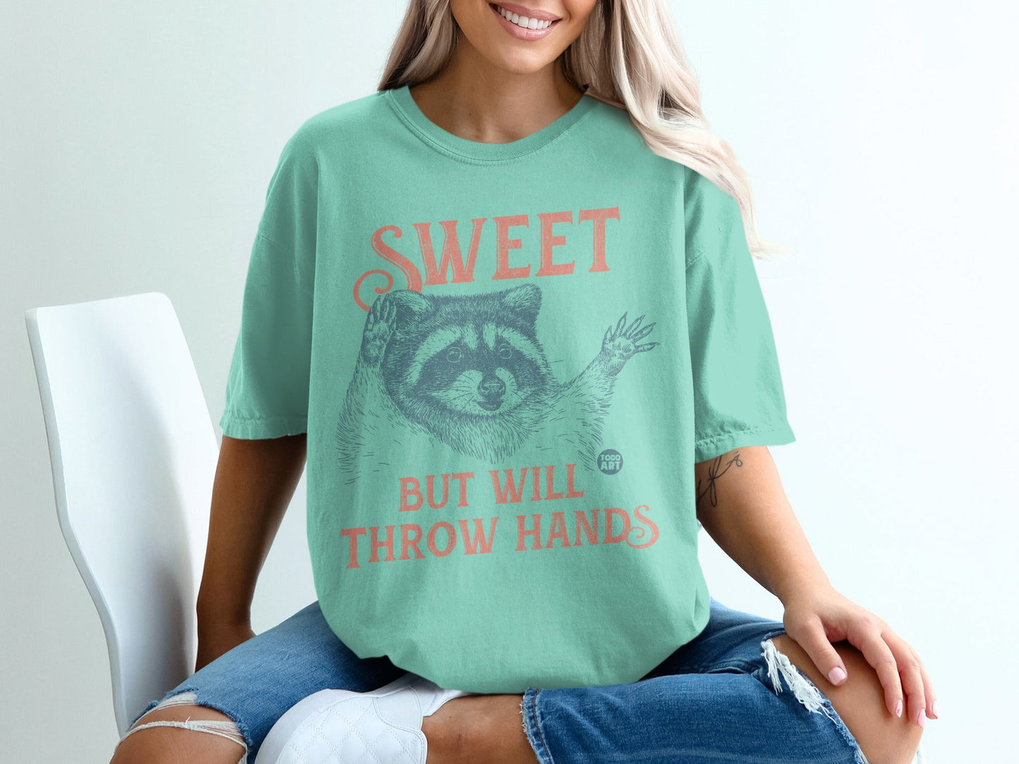 Mint t-shirt with a raccoon graphic and text "SWEET BUT WILL THROW HANDS".