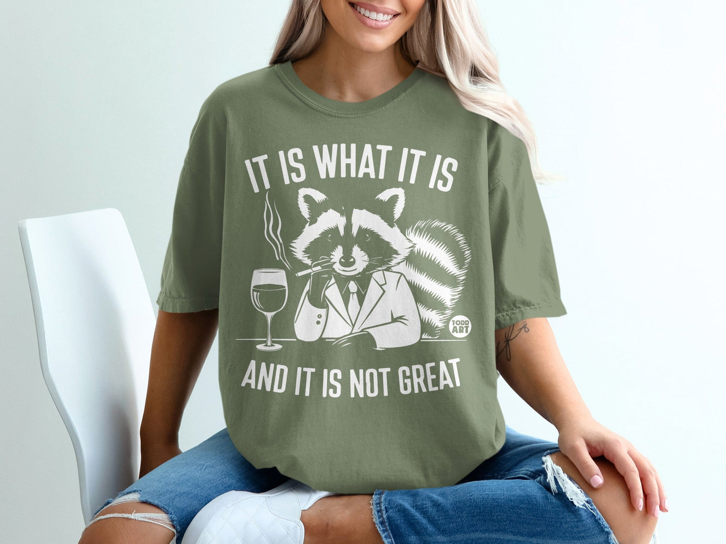 Green t-shirt with raccoon smoking and drinking, reads "It is what it is and it is not great".