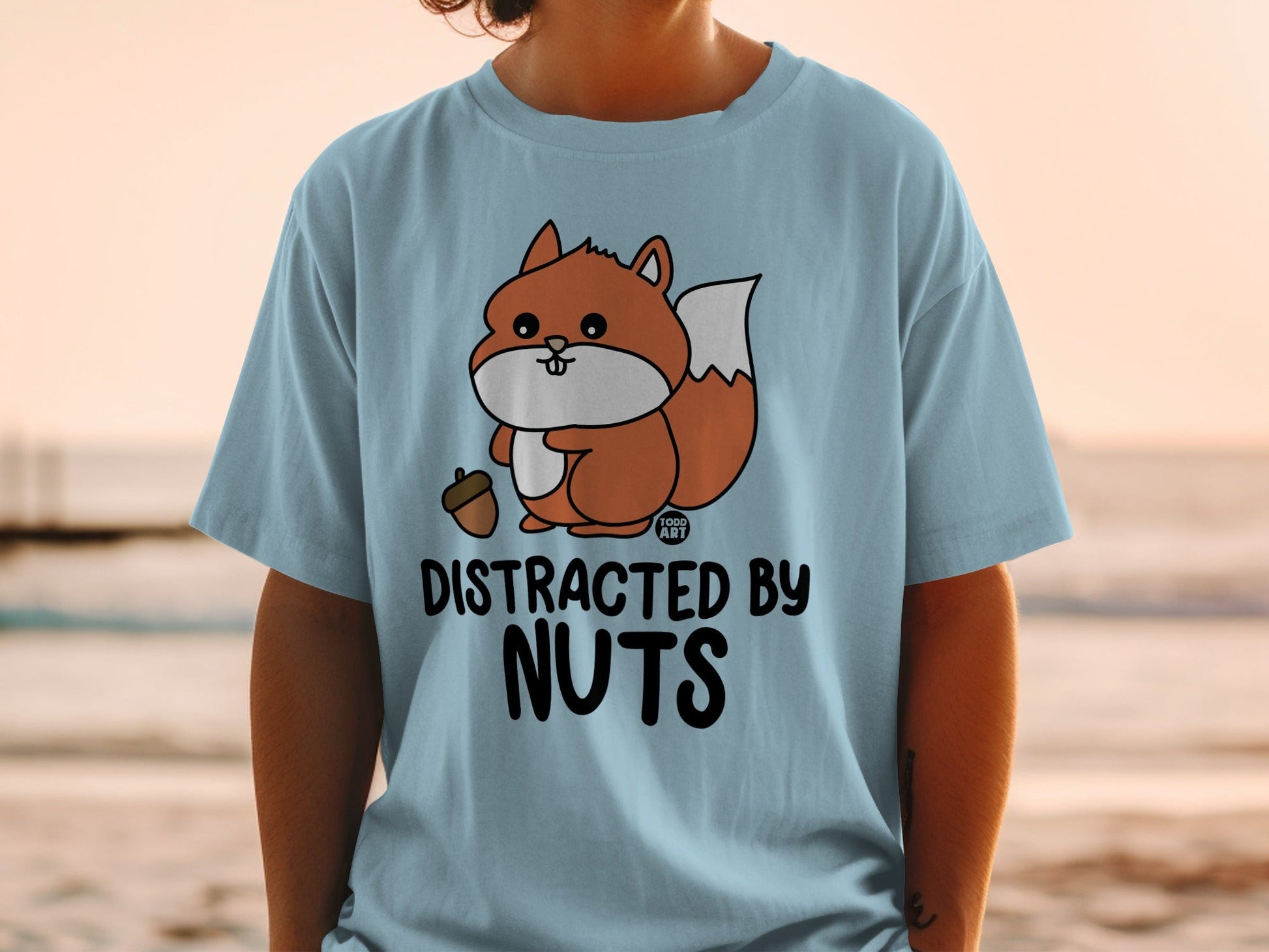 Light blue t-shirt with cartoon squirrel holding acorn and text "DISTRACTED BY NUTS".