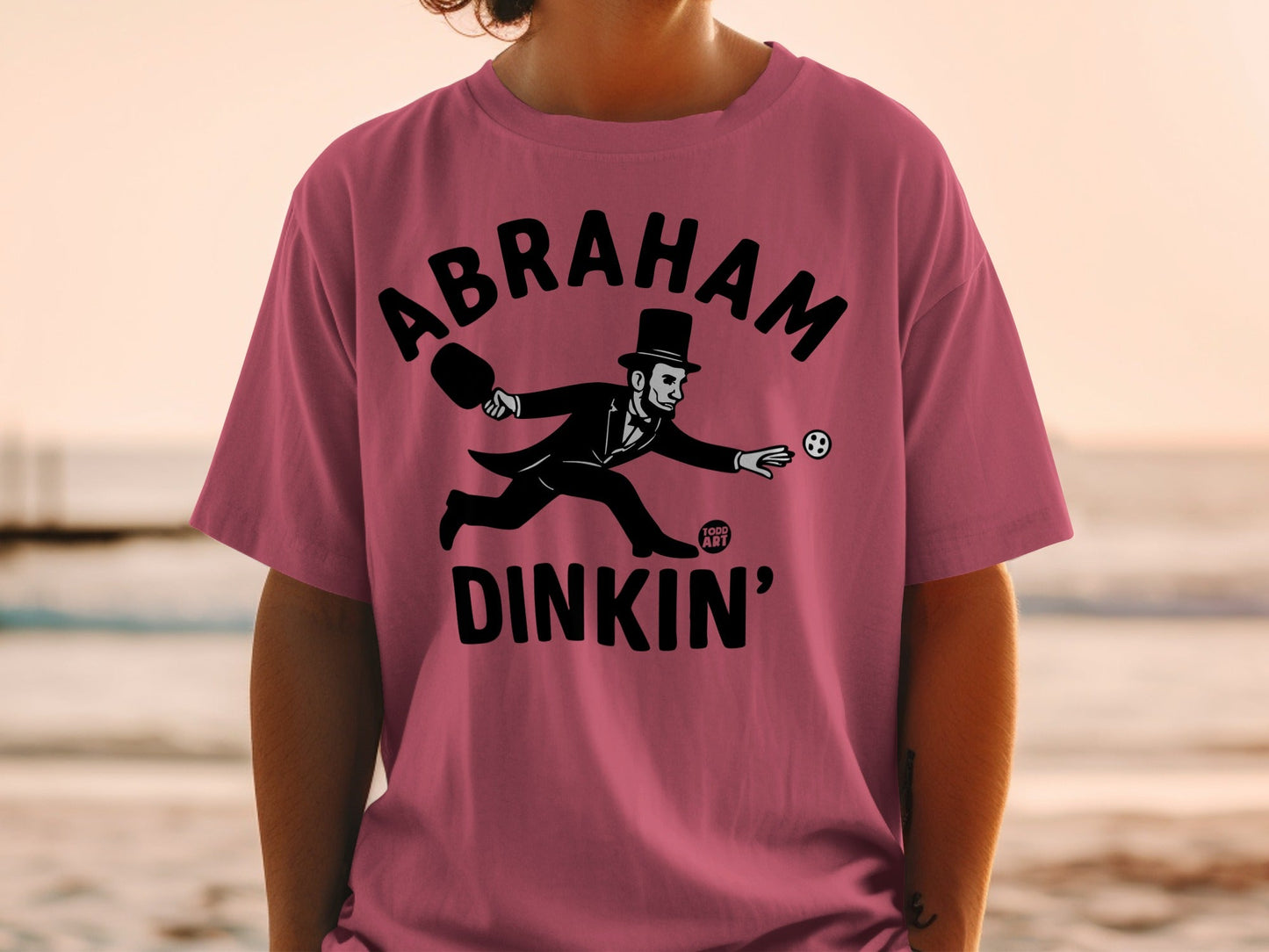 Abraham Lincoln playing pickleball on a pink t-shirt.