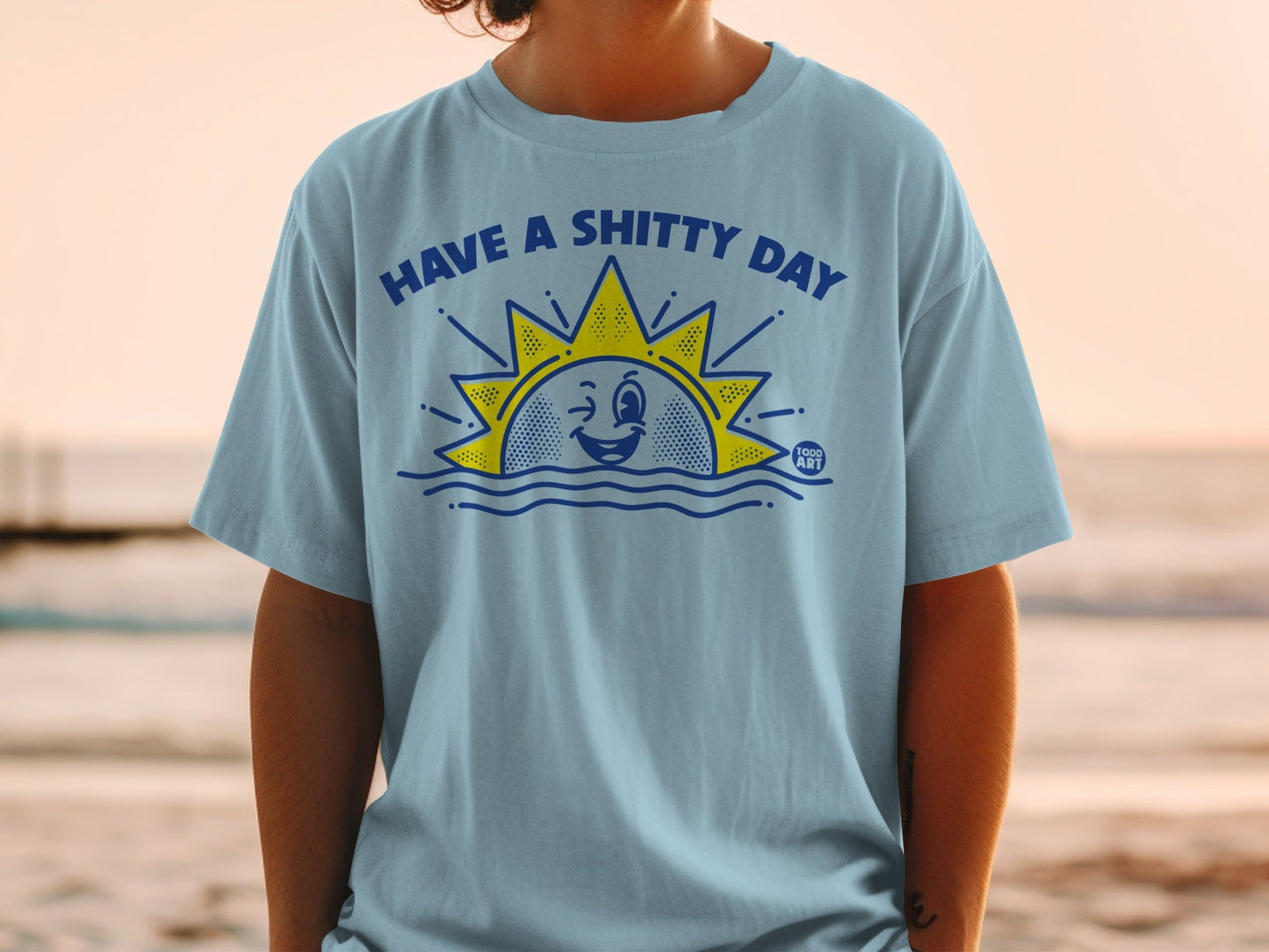 Light blue t-shirt with "HAVE A SHITTY DAY" sun graphic and "GOOD ART" logo.