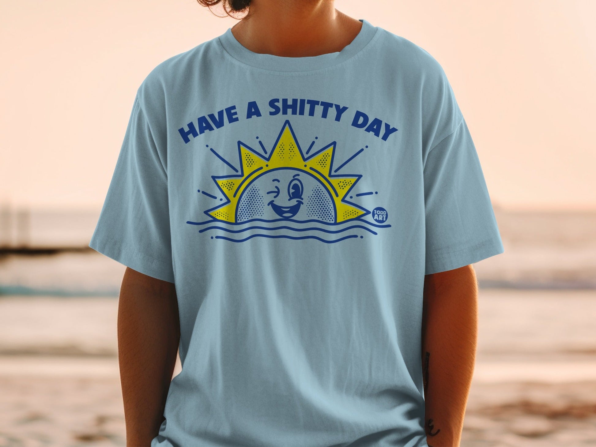 Light blue t-shirt with "HAVE A SHITTY DAY" sun graphic and "GOOD ART" logo.