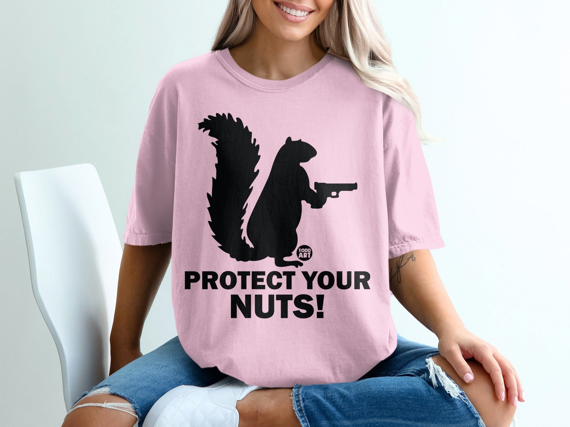 Pink t-shirt with black squirrel holding a gun and "PROTECT YOUR NUTS!" text.