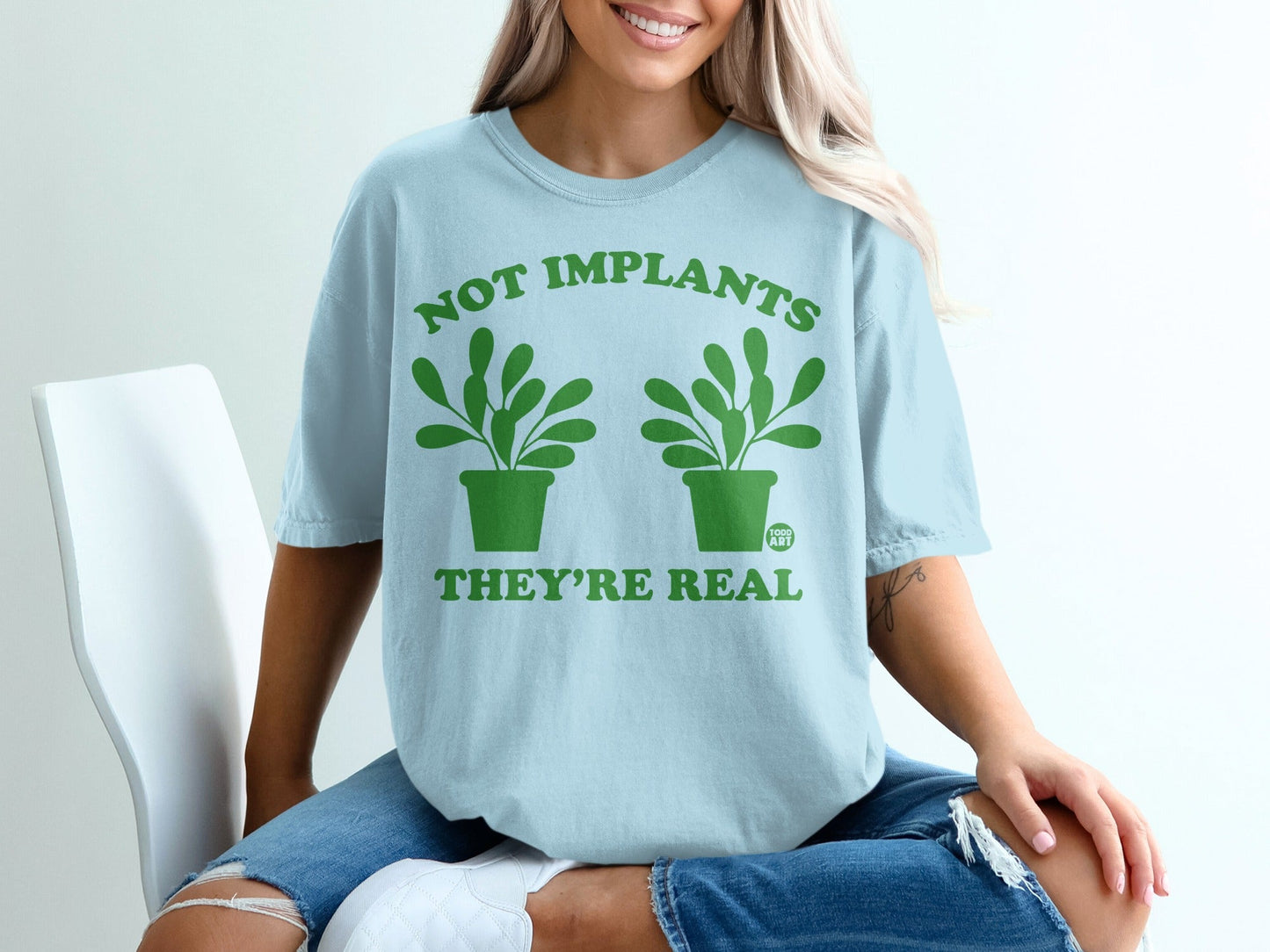 Light blue t-shirt with green plants and text "NOT IMPLANTS THEY'RE REAL".