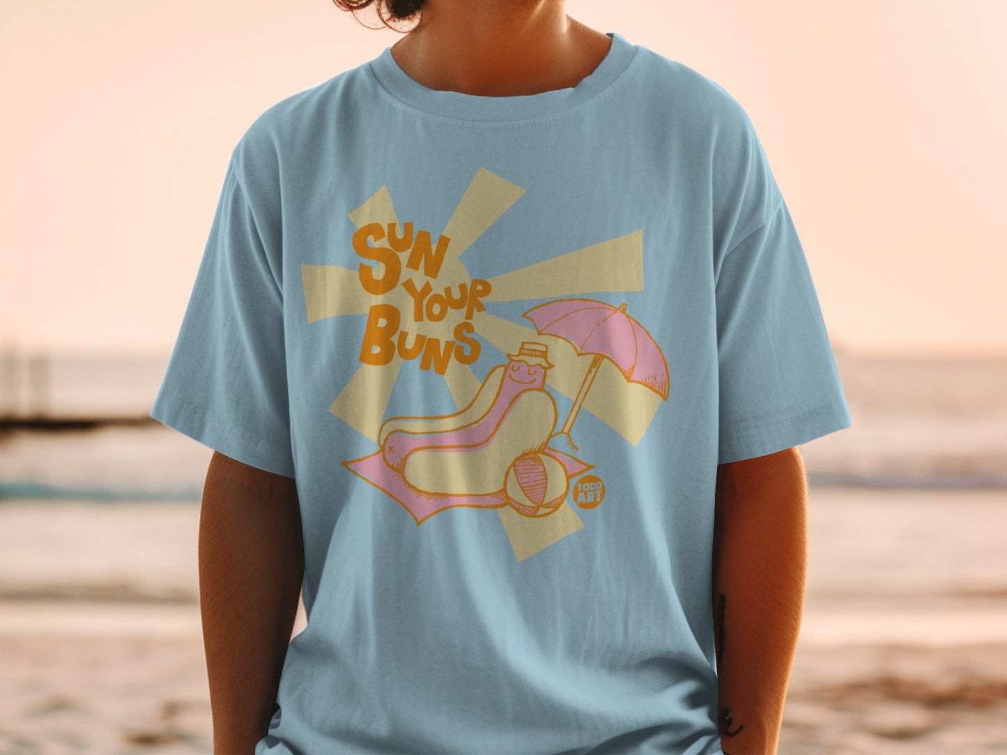 Light blue t-shirt with cartoon hot dog under umbrella on beach towel.