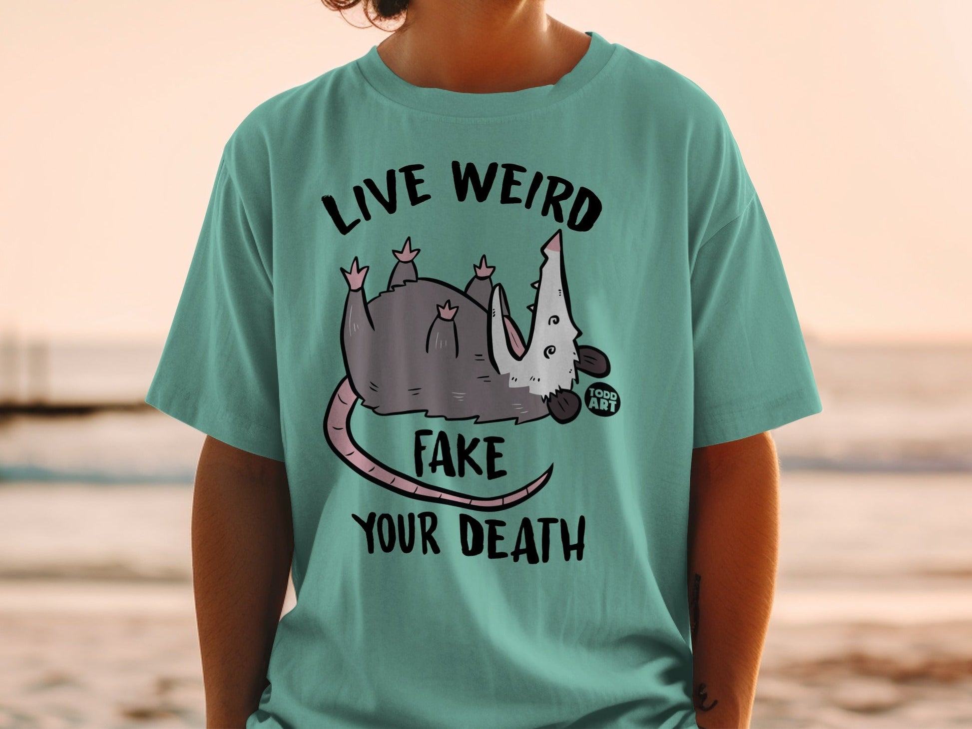 T-shirt with a cartoon opossum and text "LIVE WEIRD FAKE YOUR DEATH".