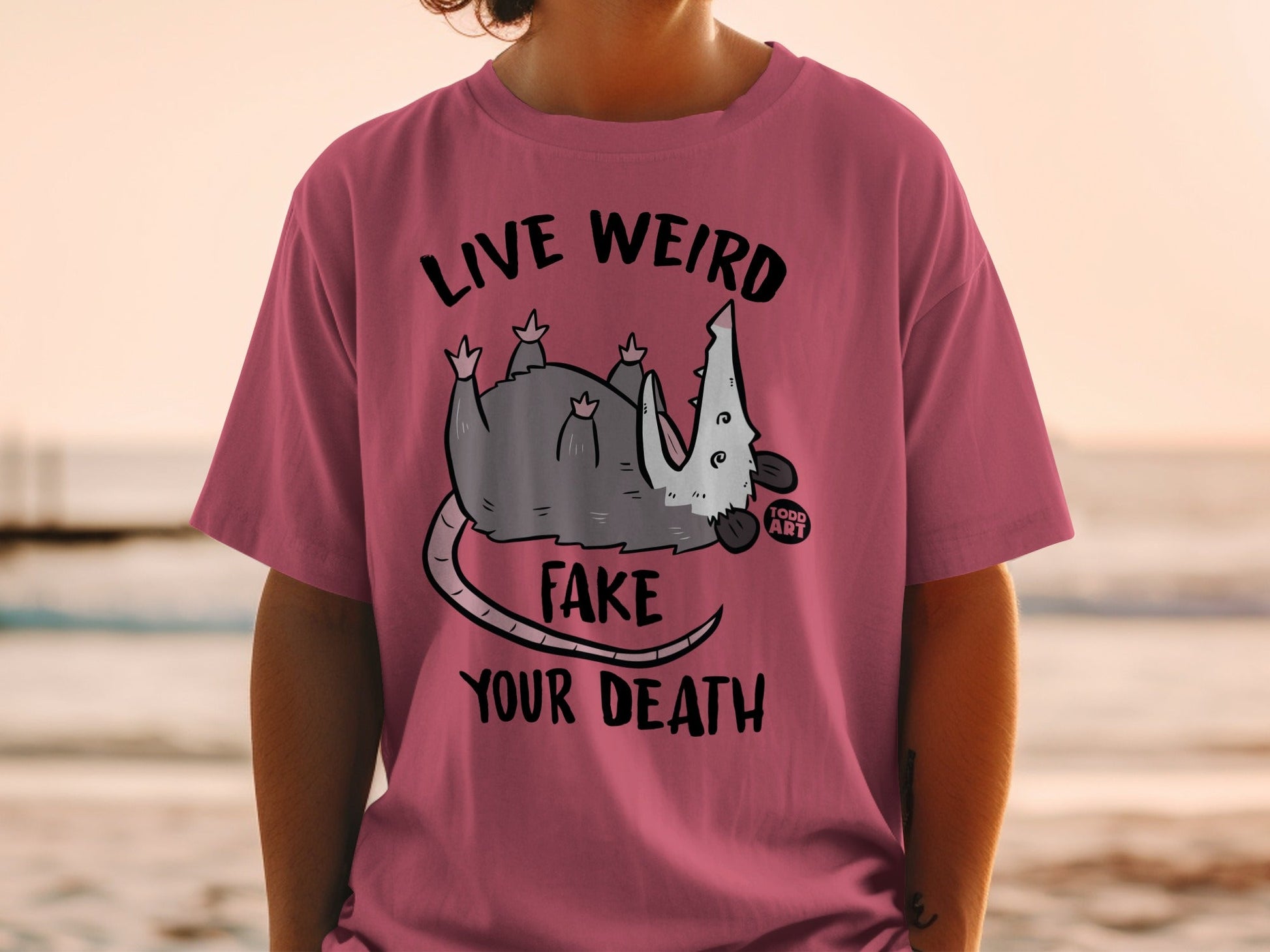 Pink t-shirt with a possum graphic and text "LIVE WEIRD FAKE YOUR DEATH".