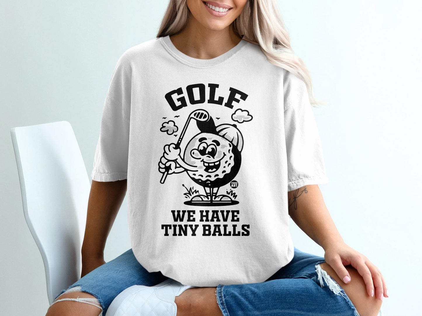 White t-shirt with black cartoon golf ball character saying "GOLF WE HAVE TINY BALLS."