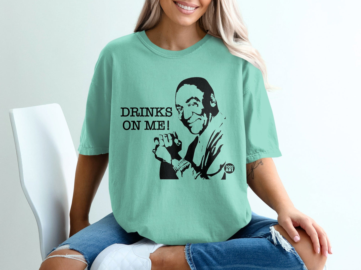 Teal t-shirt with a black graphic of a smiling man and the text "DRINKS ON ME!".
