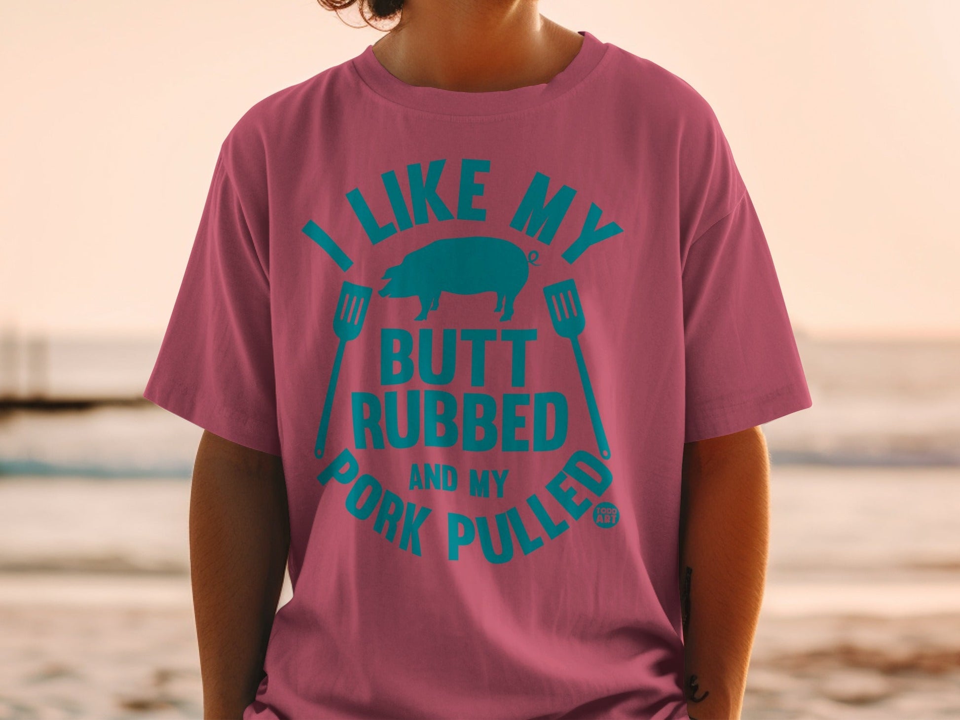 Pink t-shirt with teal text "I like my butt rubbed and my pork pulled" and pig graphic.