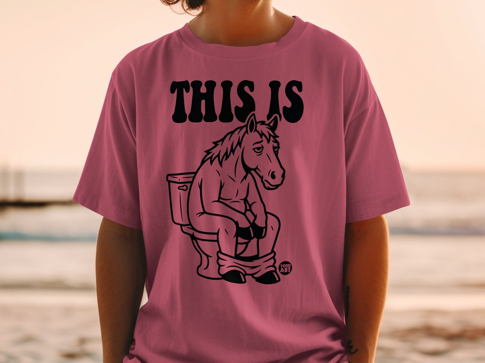Pink t-shirt with black graphic of a horse sitting on a toilet.