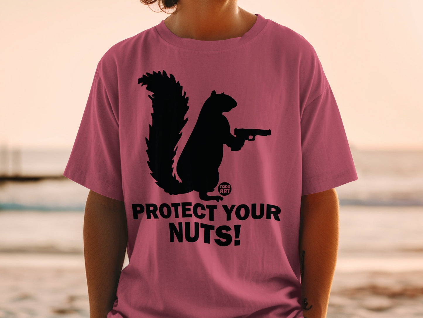 Pink t-shirt with black squirrel holding a gun and the text "PROTECT YOUR NUTS!".
