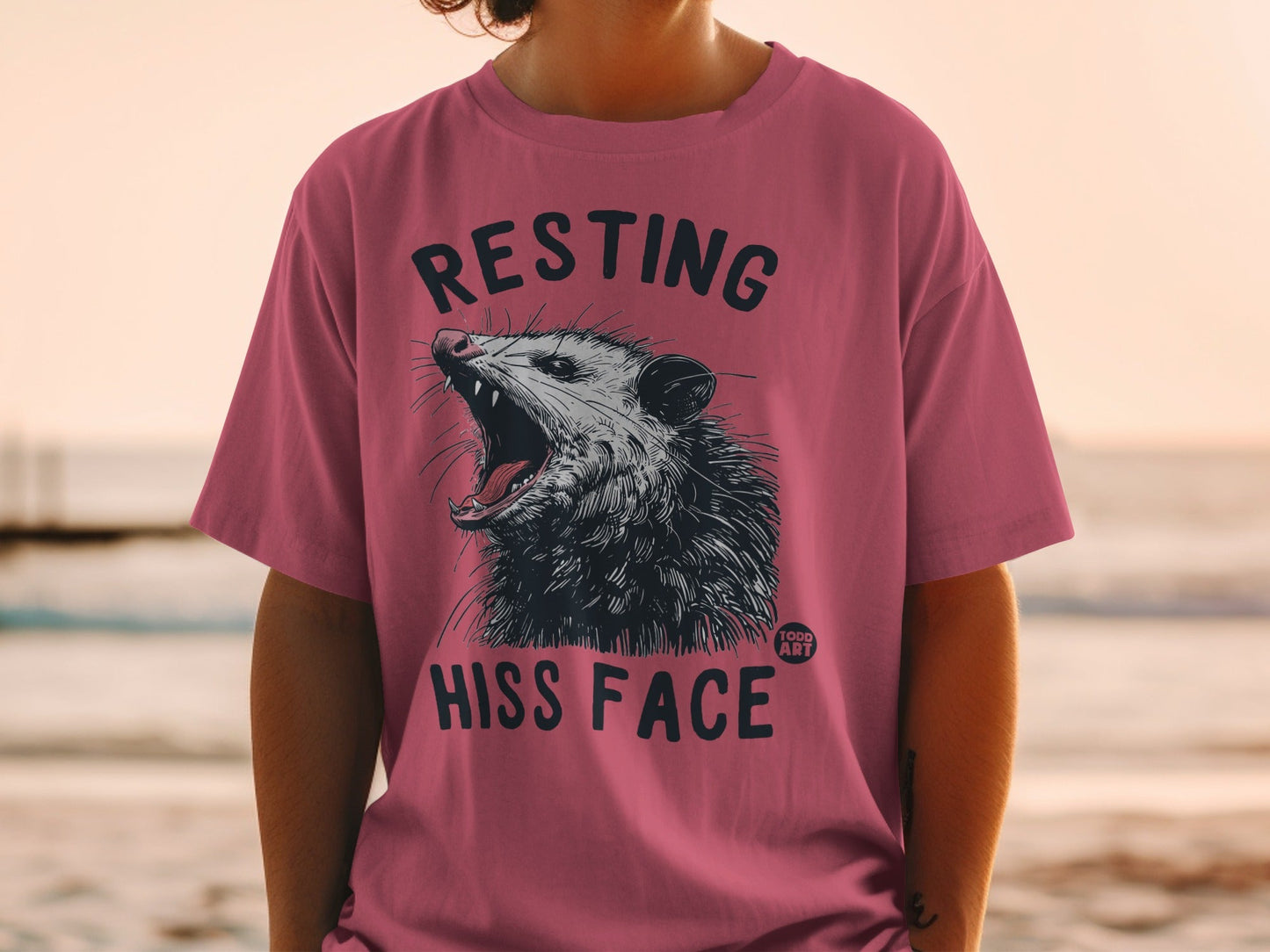 Pink t-shirt with opossum graphic and text "RESTING HISS FACE".