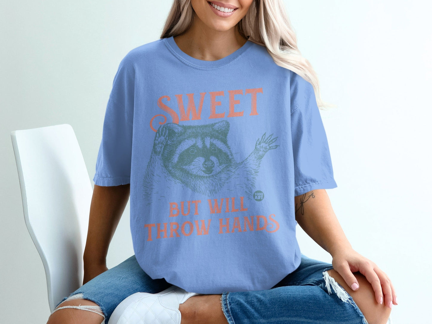 Blue t-shirt with raccoon illustration and "SWEET BUT WILL THROW HANDS" text.