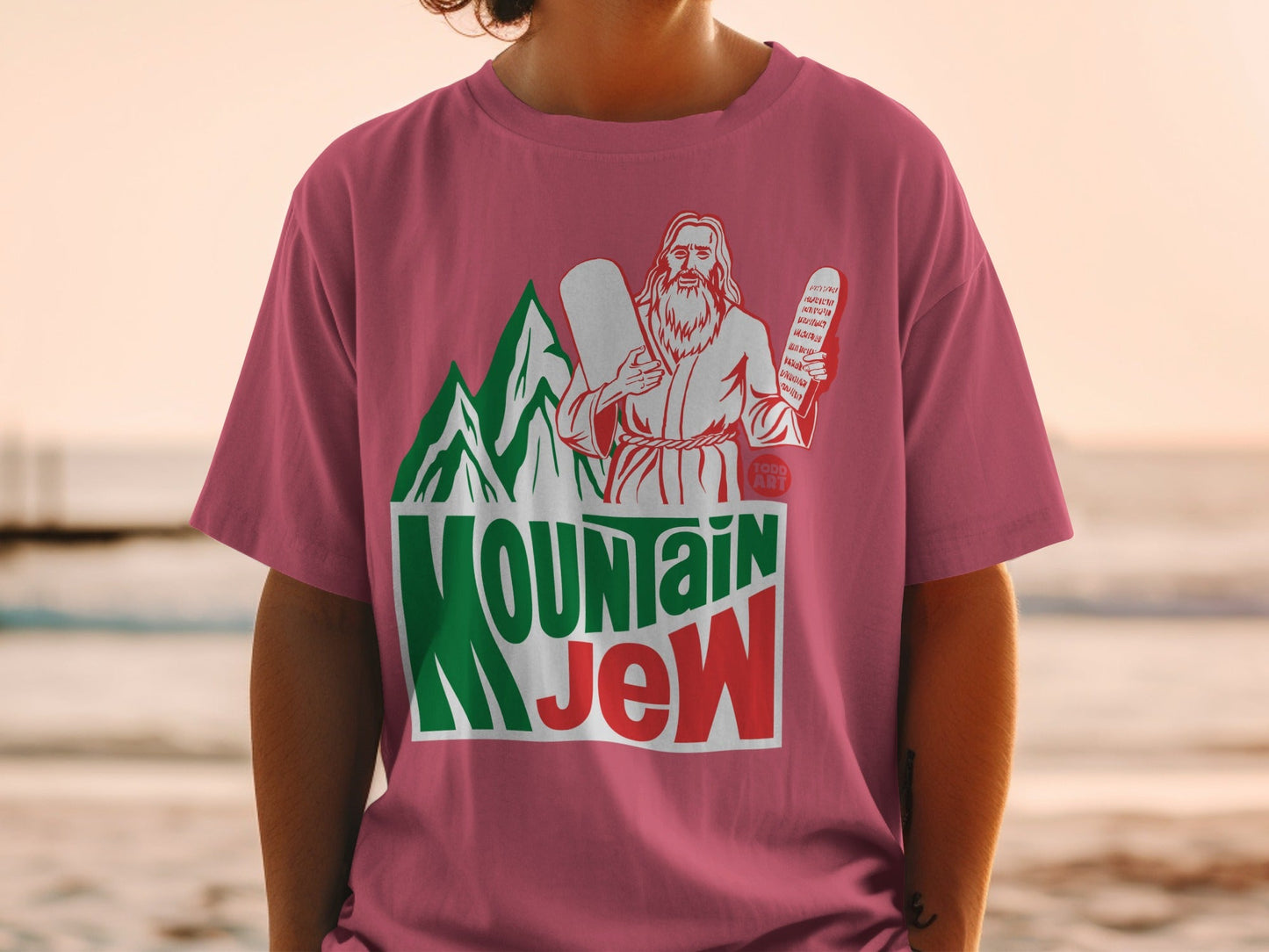 Man wearing pink t-shirt with "Mountain Jew" text, green mountains, and biblical figures.