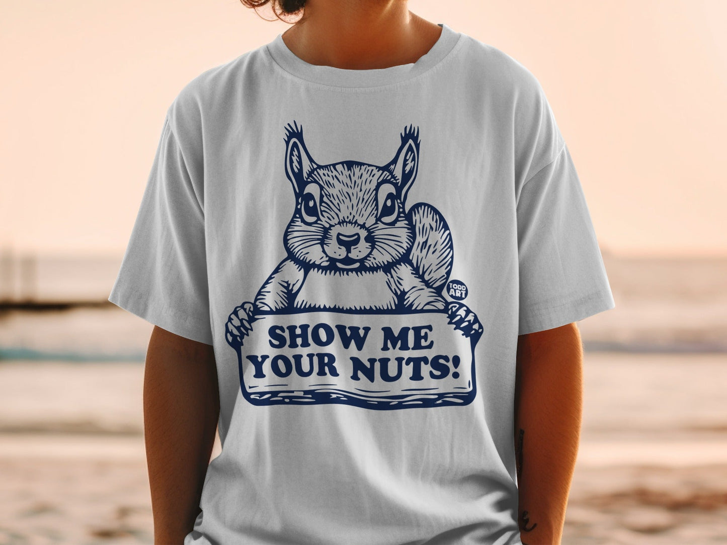 White t-shirt with a cartoon squirrel holding a sign that says "SHOW ME YOUR NUTS!"