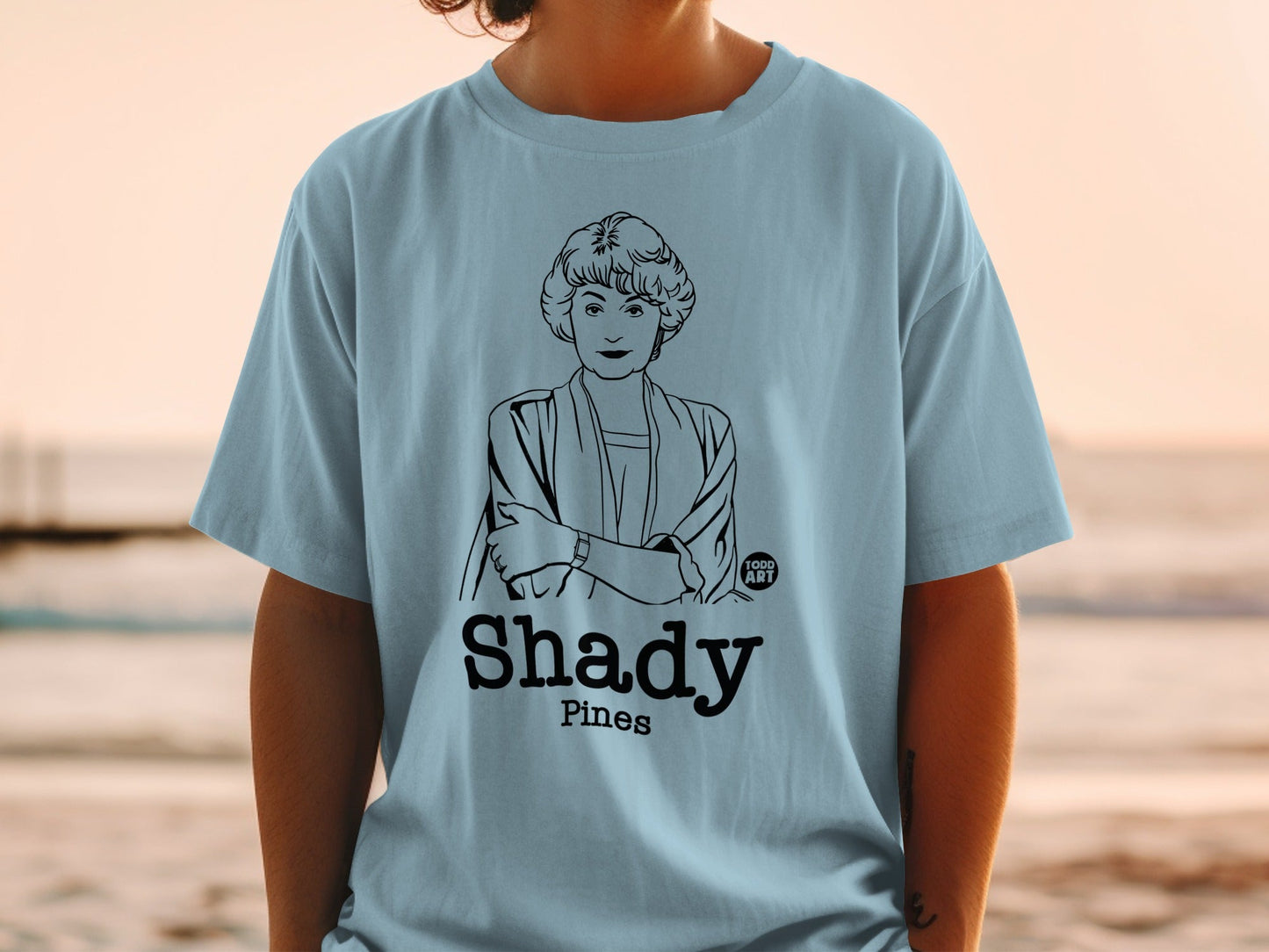 Light blue t-shirt with a black line drawing of a woman and "Shady Pines" text.