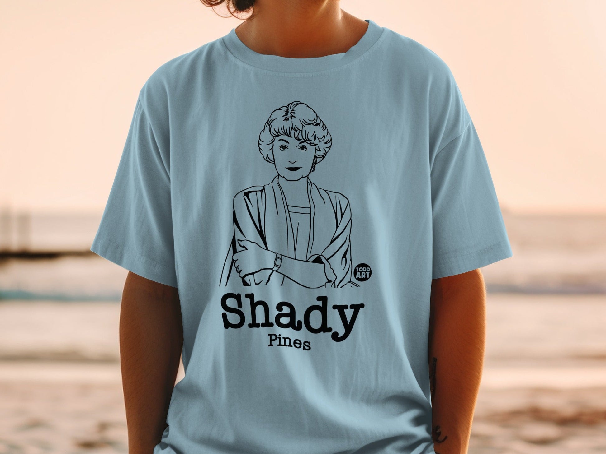 Light blue t-shirt with a black line drawing of a woman and "Shady Pines" text.