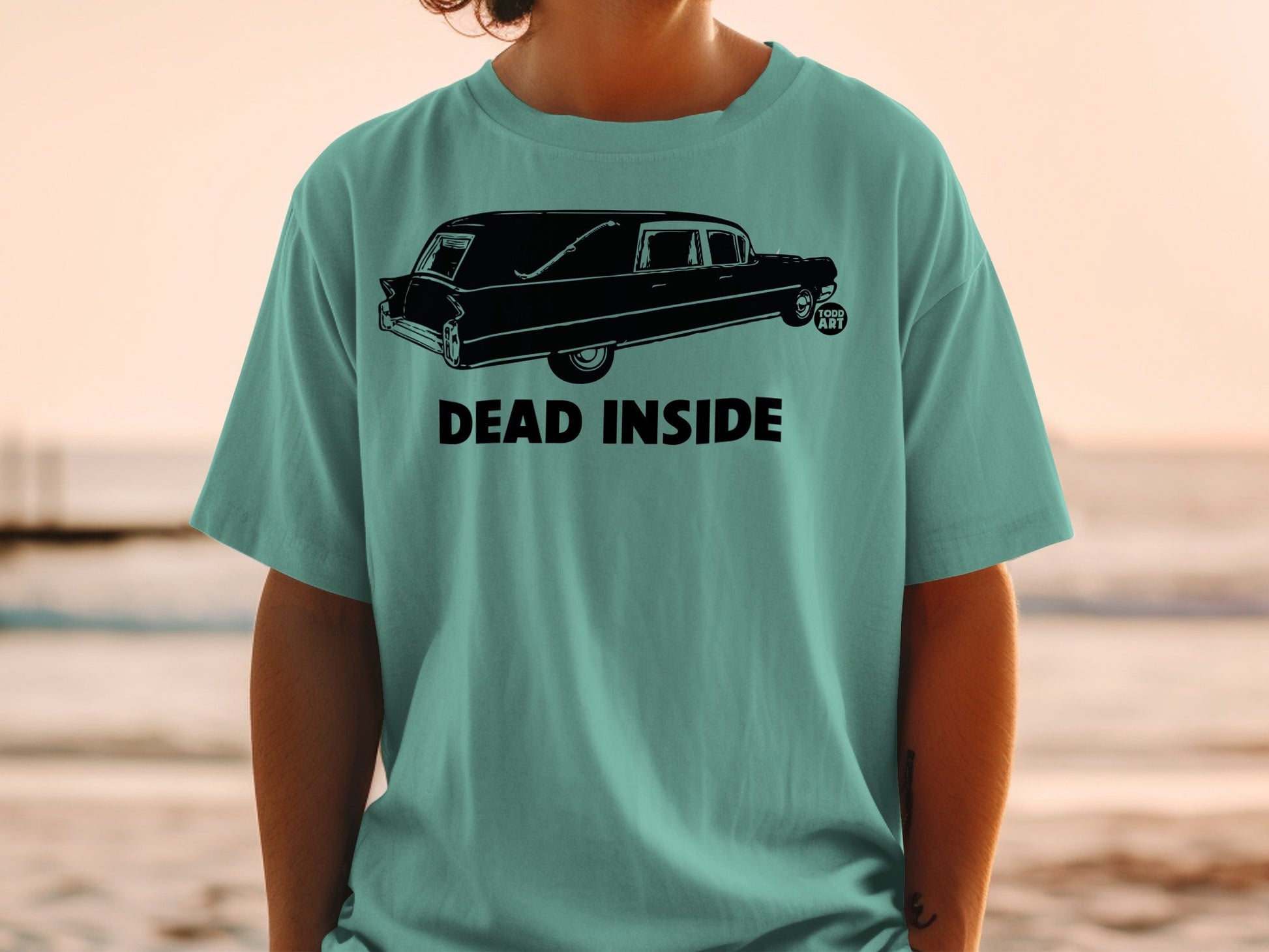 Black hearse graphic with "DEAD INSIDE" text on teal t-shirt.