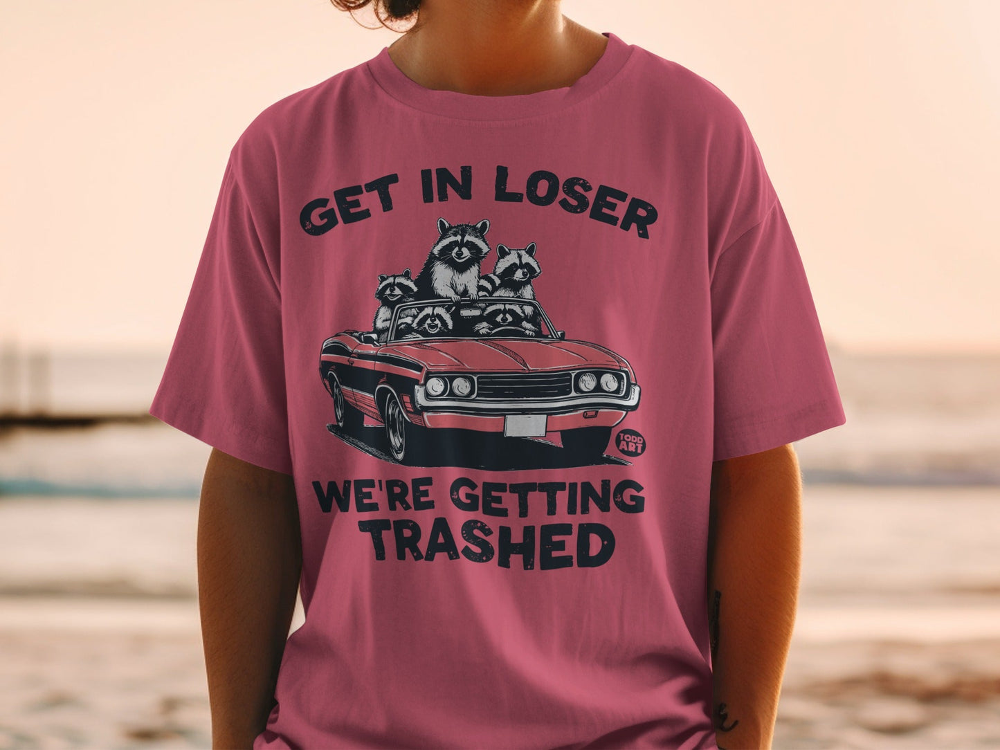 Pink t-shirt with raccoons in a convertible and the text "GET IN LOSER WE'RE GETTING TRASHED".